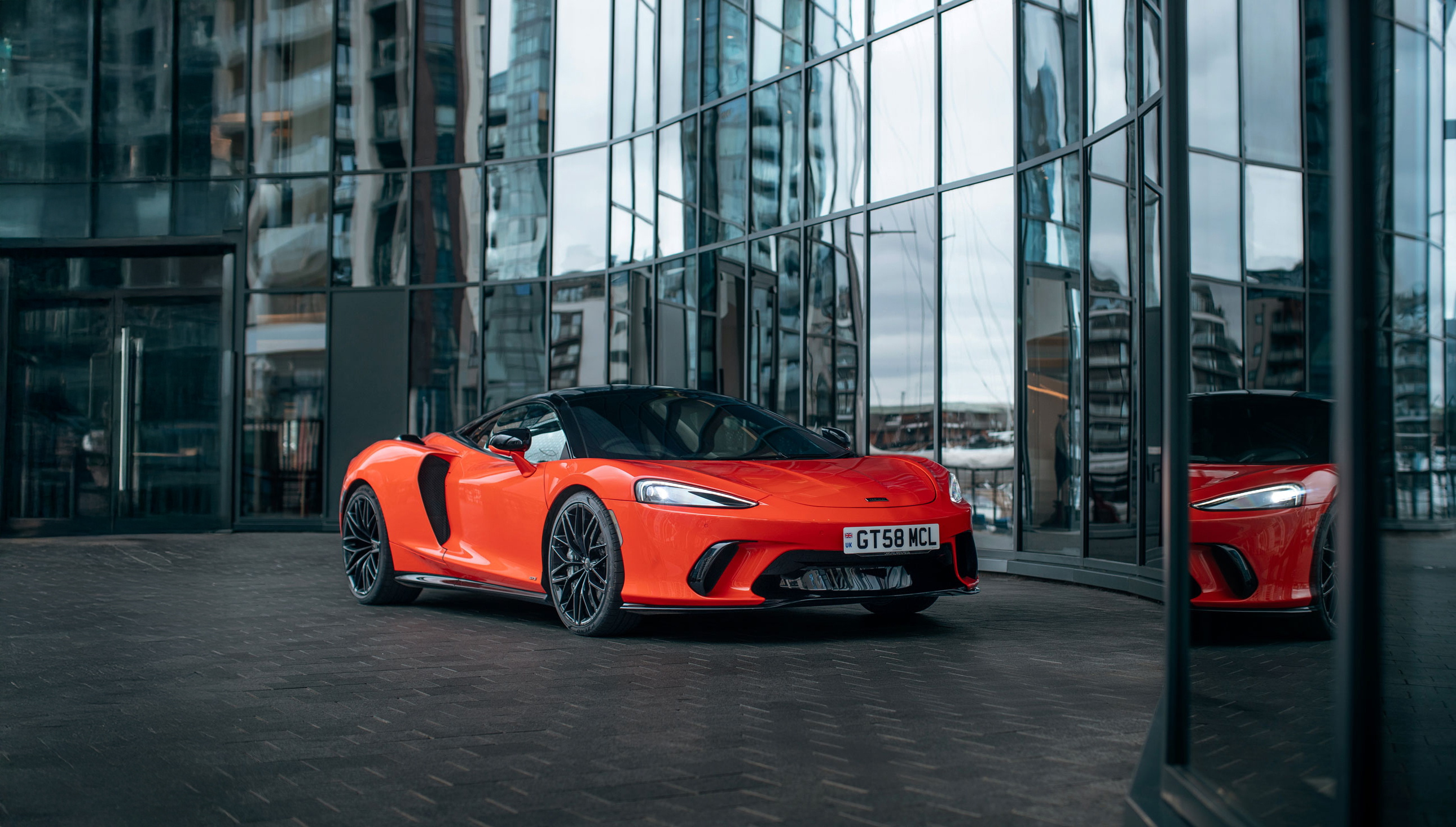 A bright red McLaren GTS is parked on a dark tiled surface in front of a modern glass building.