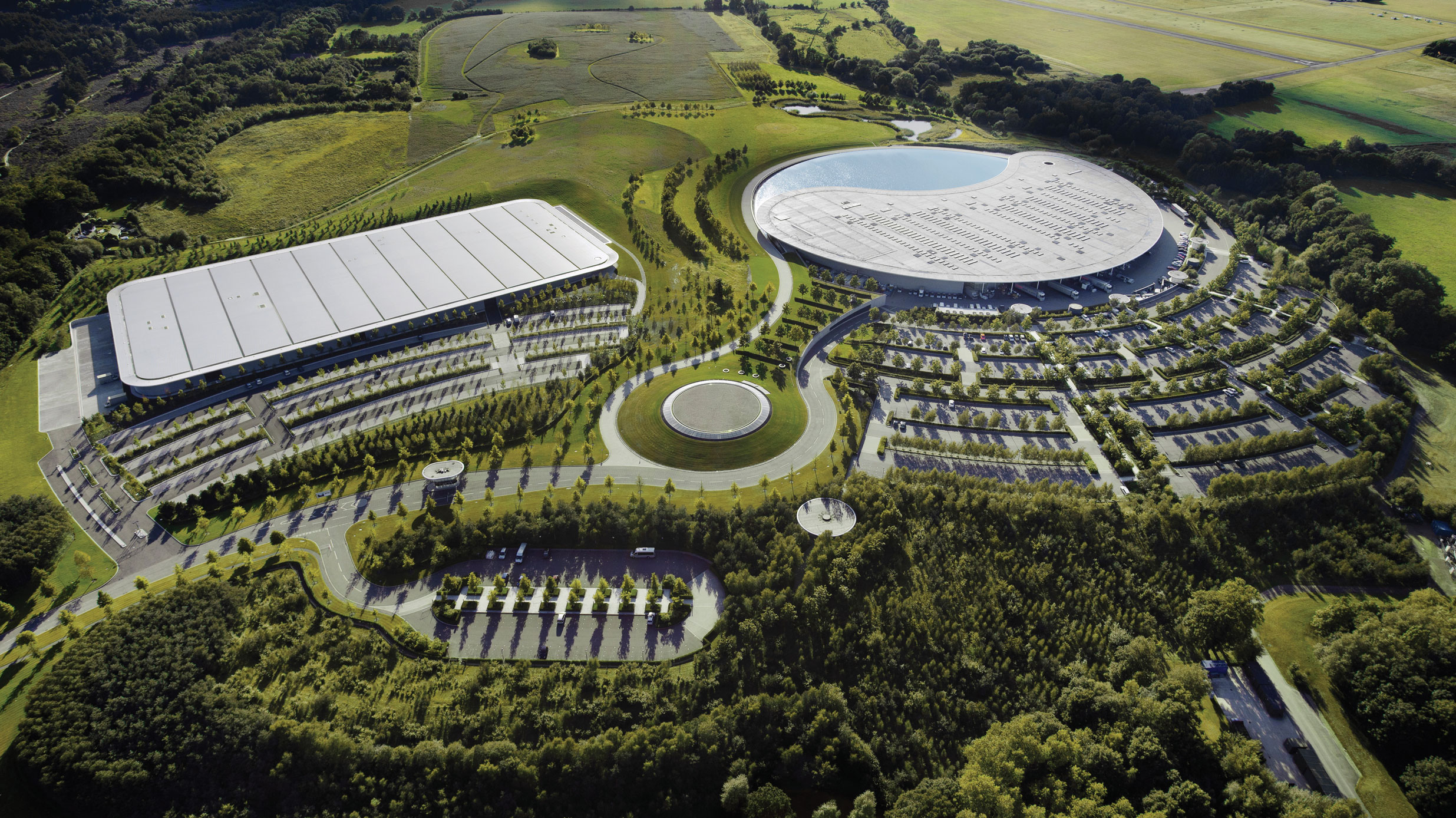 An aerial view of the McLaren Technology Centre and McLaren Production Centre in the English countryside. The image shows the main building, which is a large, curved structure with a reflective pool in the centre.