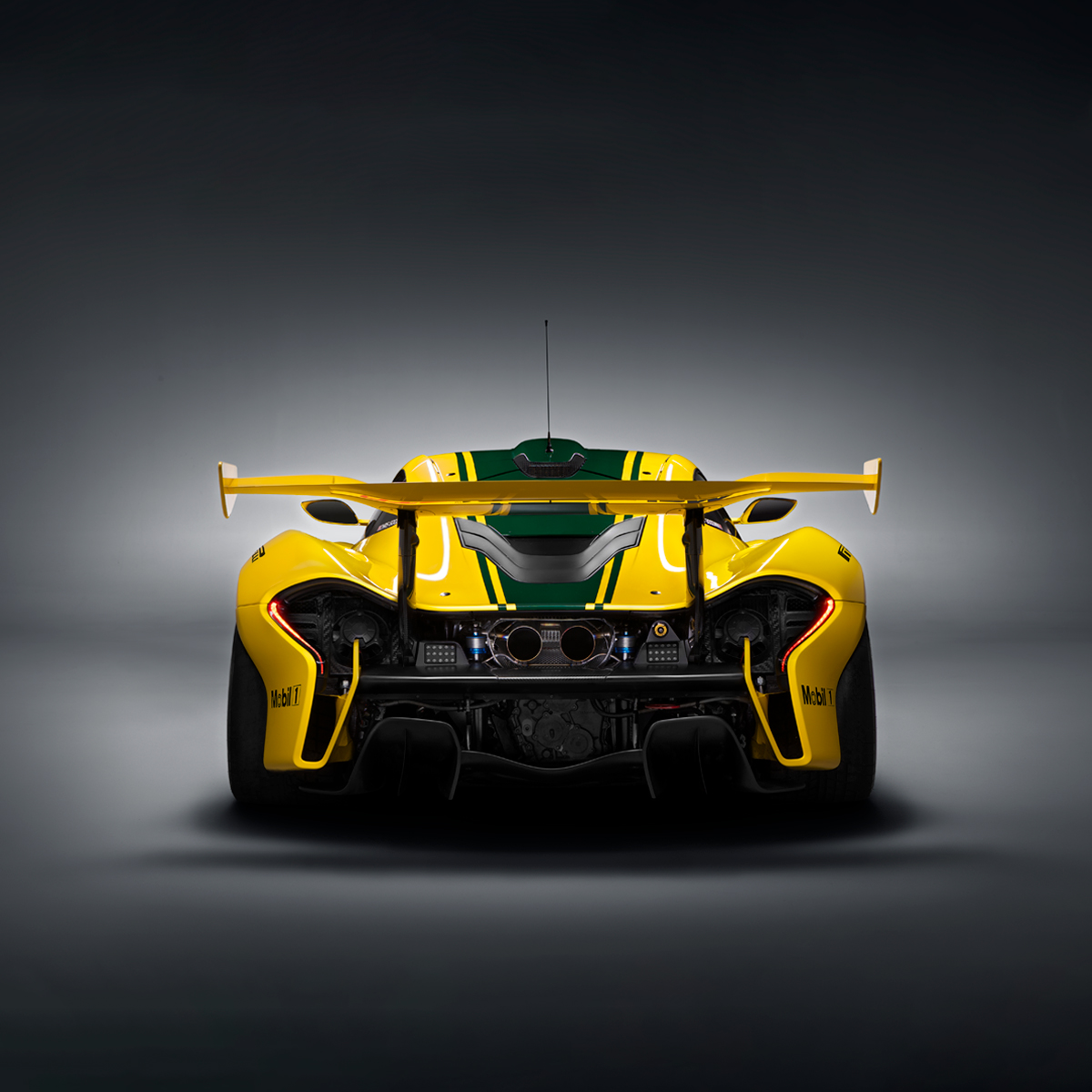 Rear view of a yellow McLaren P1 GTR with green racing stripe, number 51, and sponsorship advertising on the body, photographed in a grey studio environment.