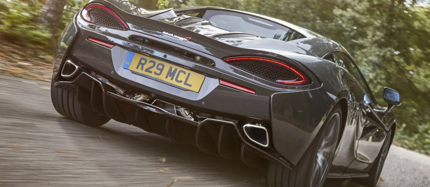  A rear low-angle shot of a dark grey McLaren supercar accelerating on a winding road, showcasing its distinctive taillights, exhaust pipes, and license plate, with sun-dappled trees in the background.