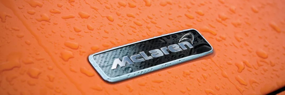 Detail shot of a silver and black McLaren brand badge against a vibrant orange, wet background.