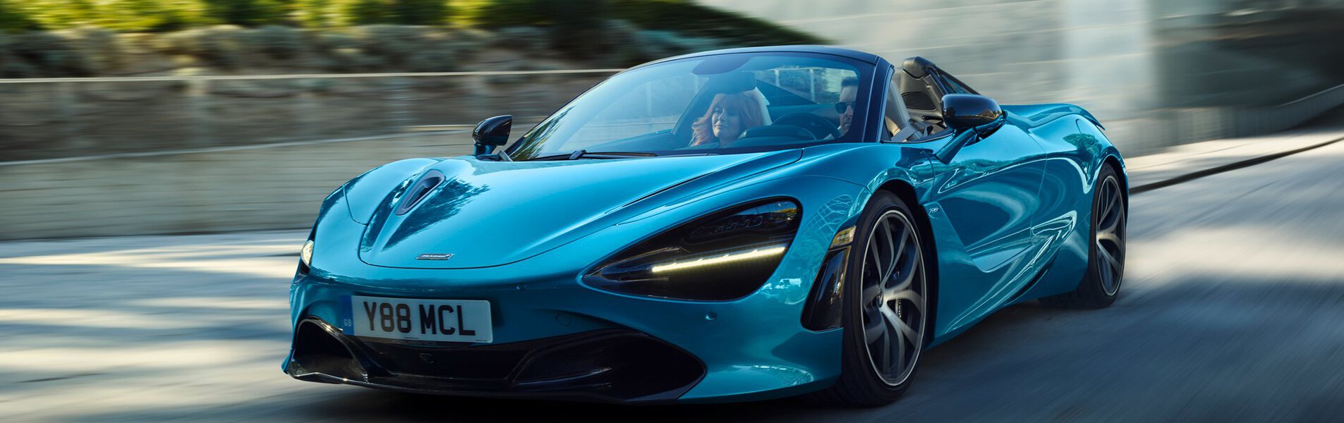 A vibrant blue McLaren 720S Spider convertible is driving towards the camera on a road, with motion blur in the background suggesting speed.