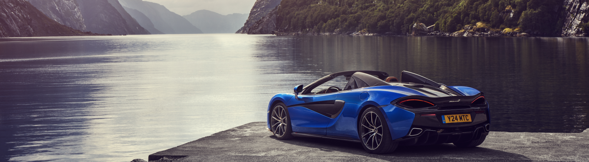 A blue McLaren 570S Spider convertible is parked by a scenic lake with a large mountain or cliffside in the background, showcasing its side profile.
