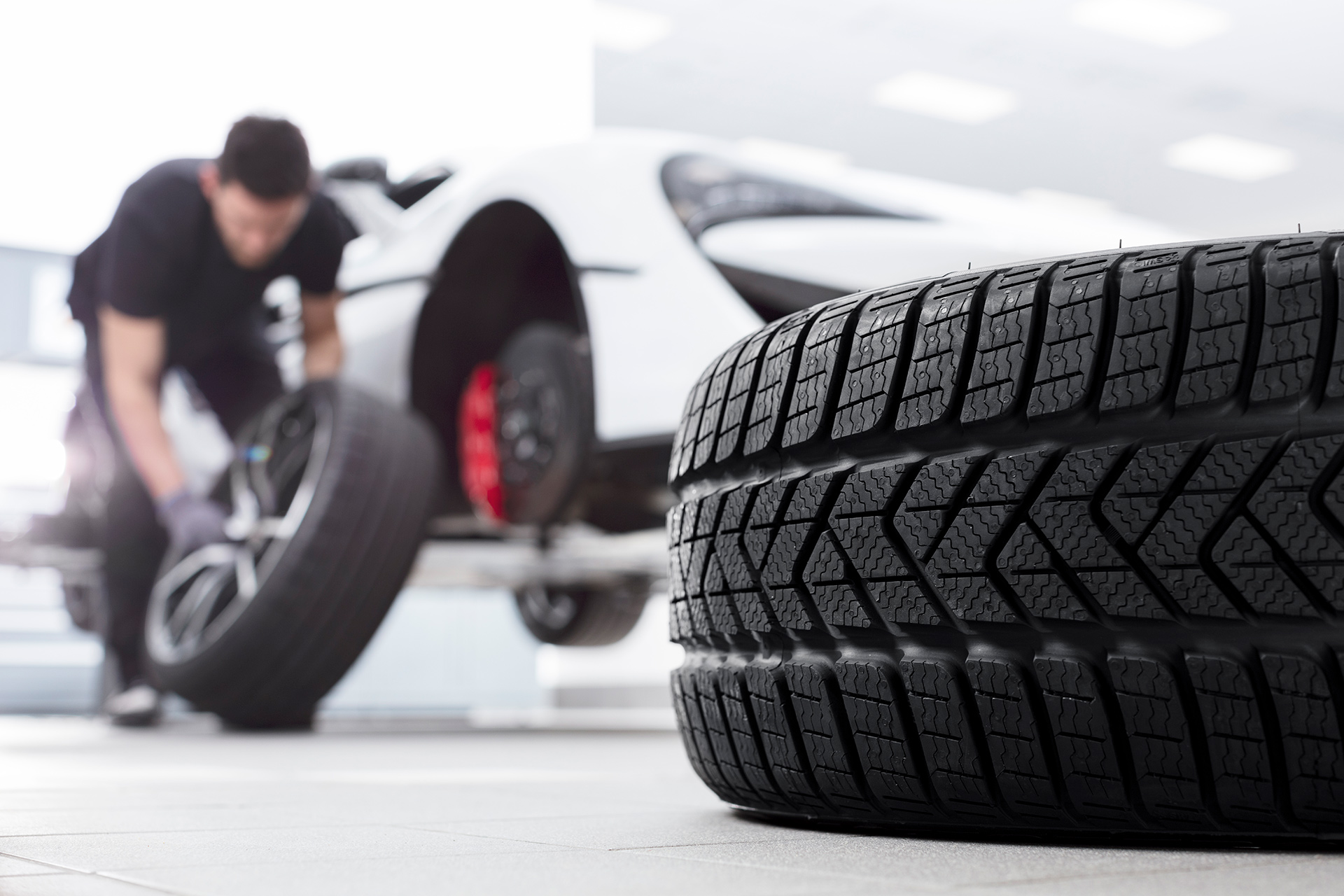 A large tire in the foreground with a McLaren technician changing a wheel on a white supercar in the background.