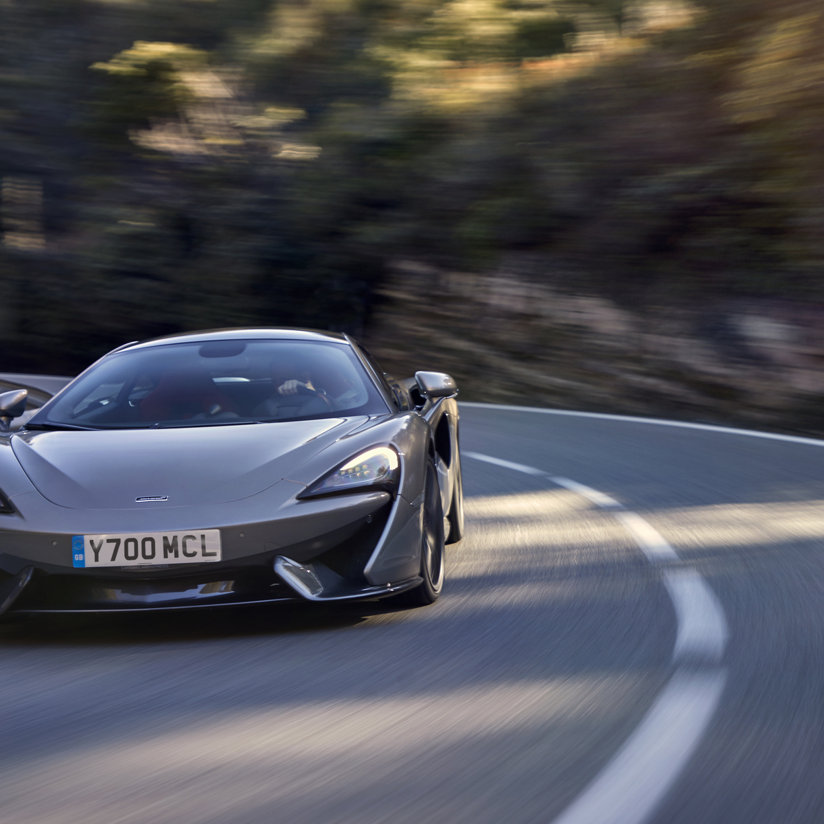 A grey McLaren 570S sports car is shown cornering on a winding road with motion blur, conveying speed and performance.