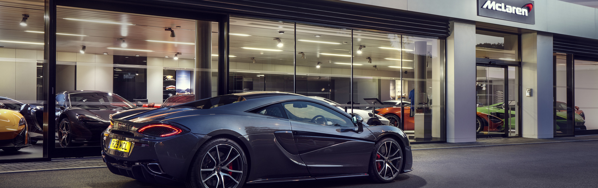 A grey McLaren 570S supercar parked outside a McLaren dealership showroom with large glass windows.