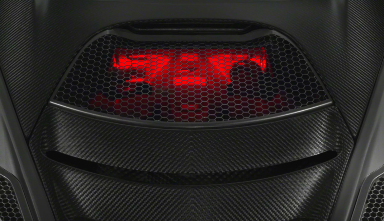 Close-up detail shot of the red, glowing light and honeycomb grille on the rear deck of a McLaren supercar, framed by carbon fiber bodywork.