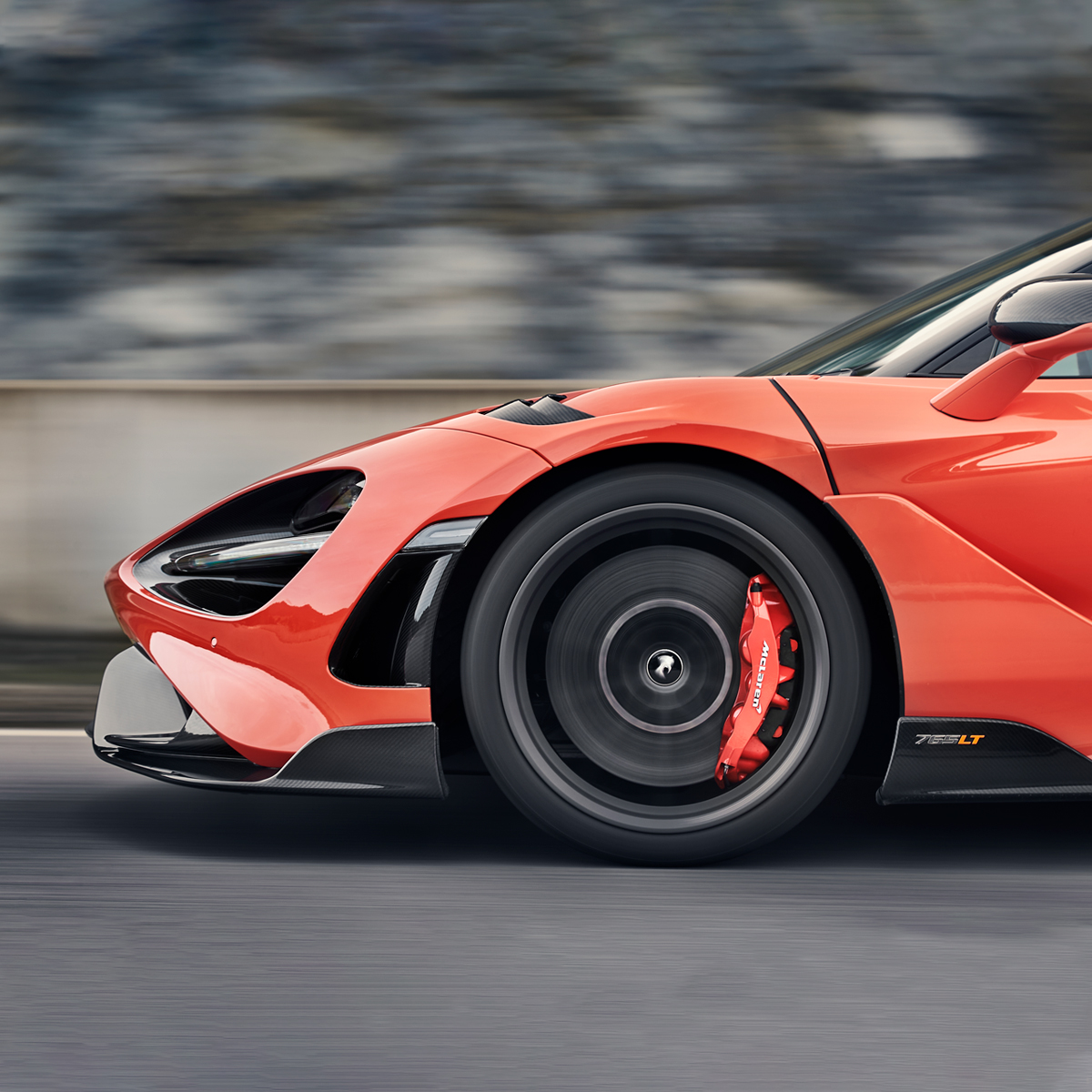 A close-up, side profile shot of a vibrant orange McLaren 765LT, showcasing the front wheel, red brake caliper, and intricate aerodynamic vents while the car is in motion.
