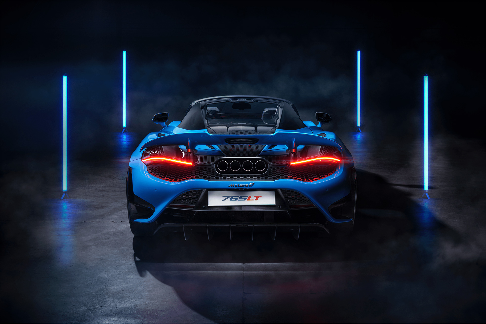 A vibrant blue McLaren 765LT Spider is shown from the rear in a dark garage, with its brake lights glowing red and two vertical blue neon light strips on either side.