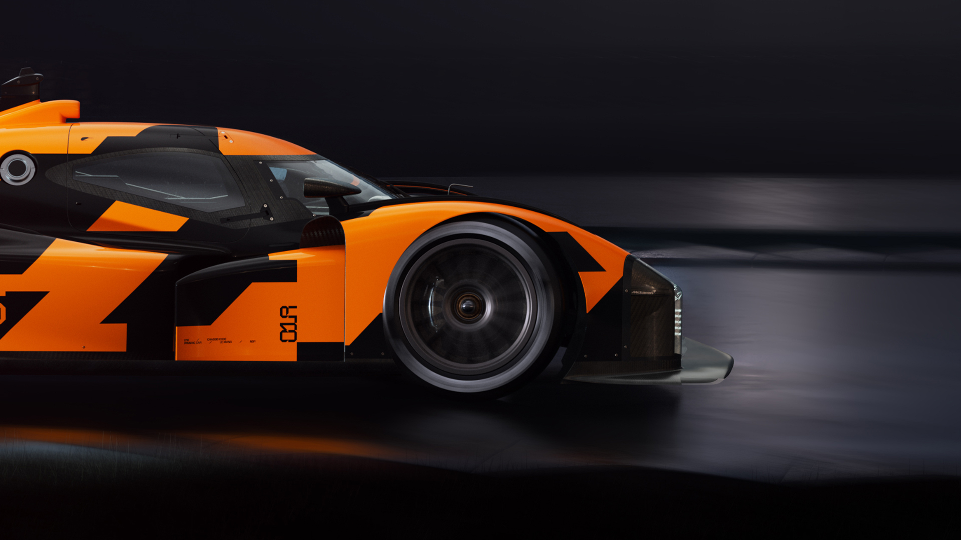 A low-angle side-profile shot of a black and orange McLaren endurance race car prototype in motion, with a motion-blurred background.