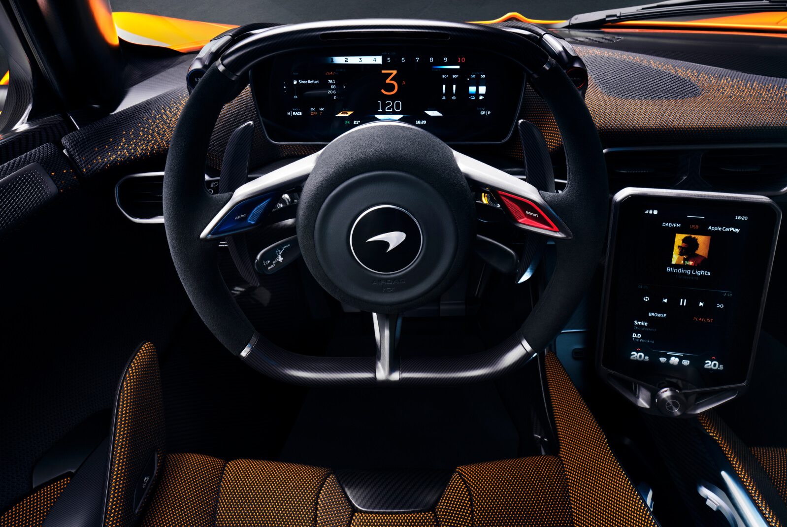 A dashboard of a McLaren W1 with a steering wheel, showing the driver display and the carbon fibre design of the steering wheel with shift lights and blue and red buttons.