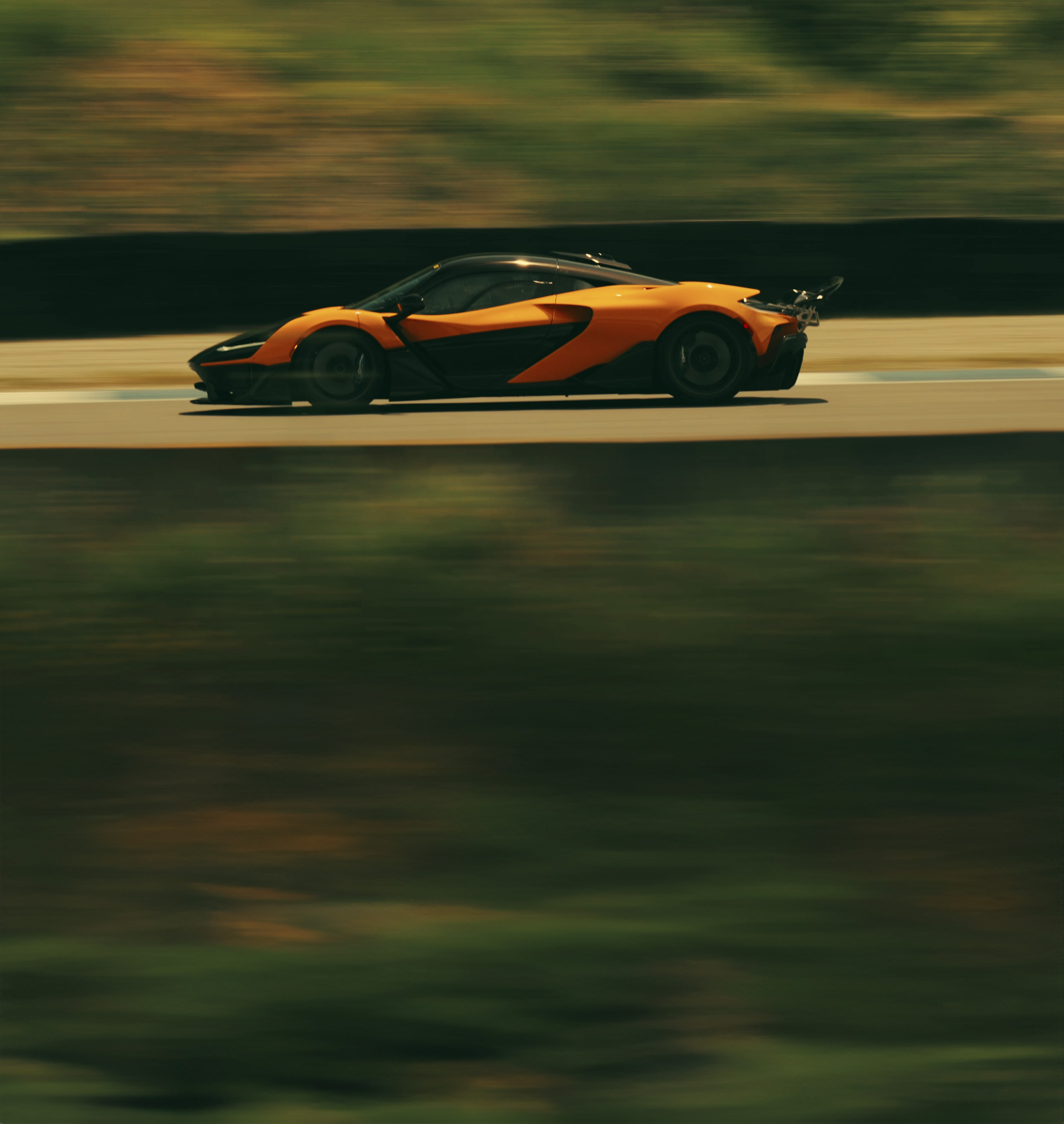 A side-on dynamic shot of an orange and black McLaren W1 speeding down a track with a motion-blurred background.