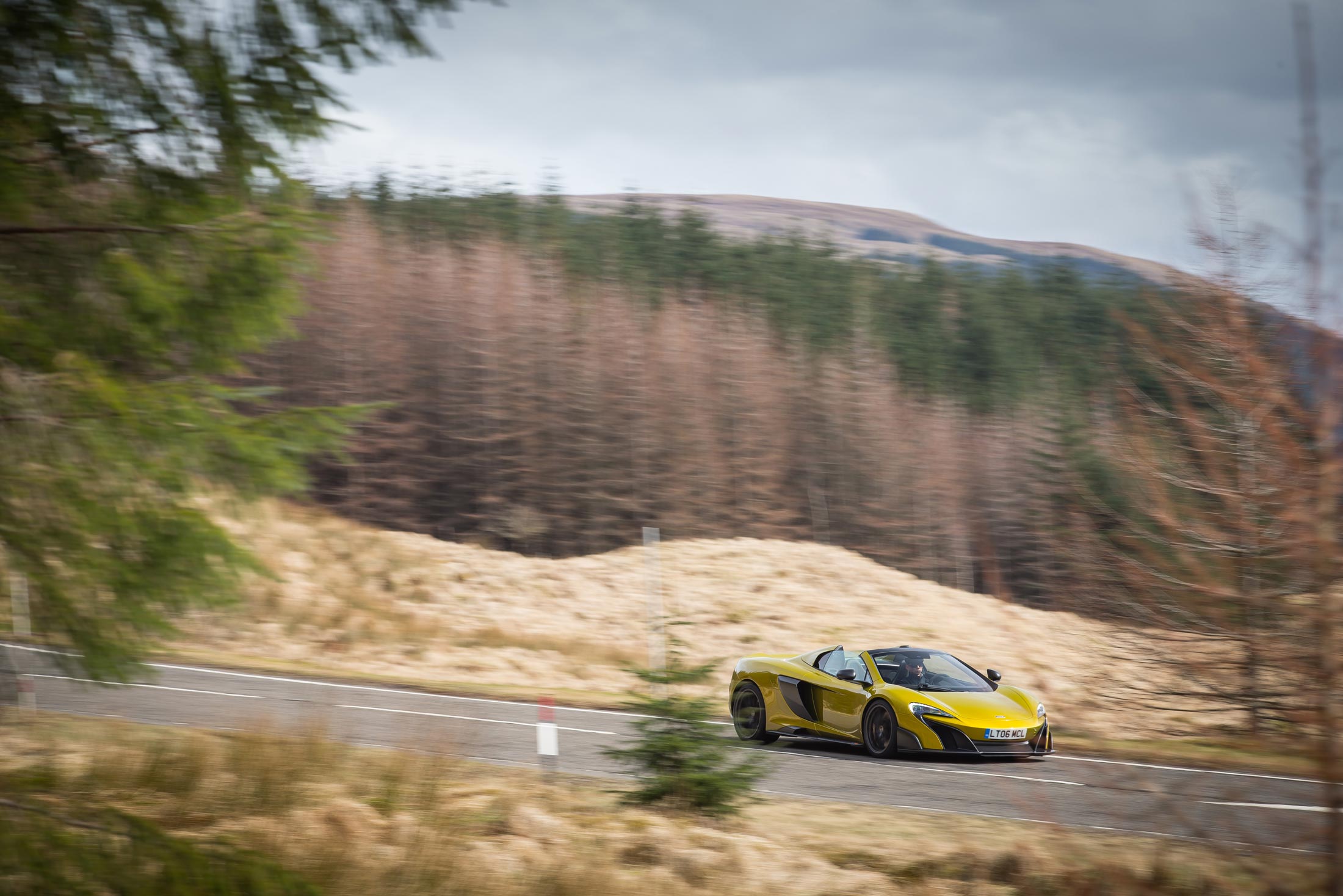 A yellow McLaren 675LT is captured in motion on a winding country road, with motion blur indicating speed and a wooded landscape in the background.