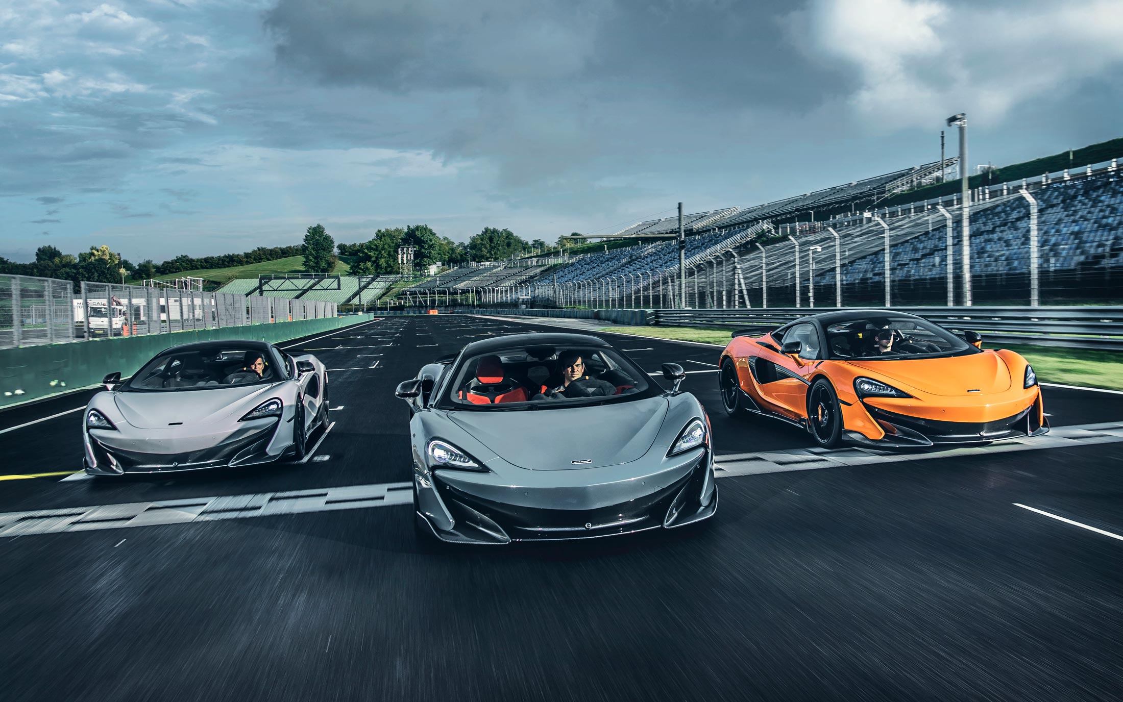 A trio of McLaren 600LTs (two grey and one orange) driving together on a racetrack, captured from the front.