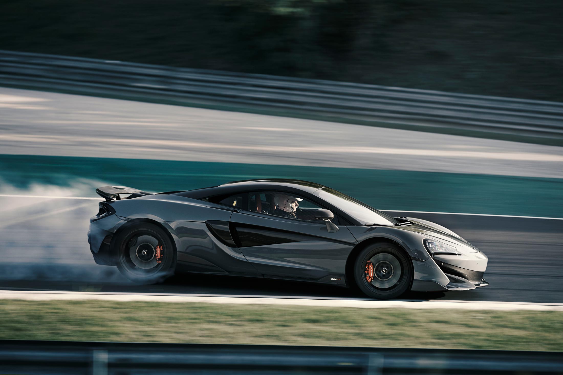 A dynamic side-profile shot of a grey McLaren 600LT on a racetrack, with a driver in a helmet visible in the cockpit and tire smoke trailing from the wheels.            Analysis