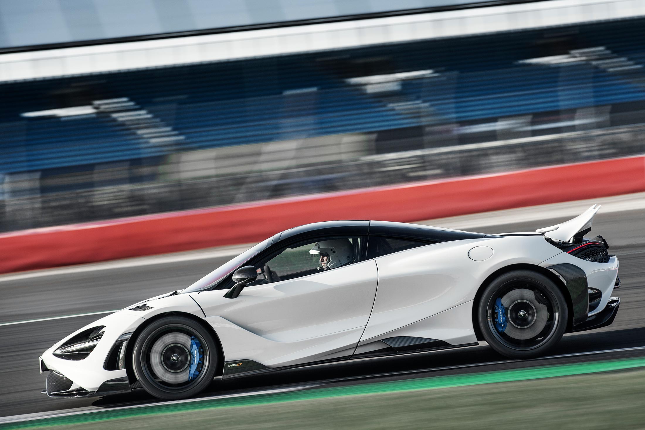 dynamic side-profile shot of a white McLaren 765LT speeding on a racetrack, with a driver in a helmet and blue brake calipers visible.