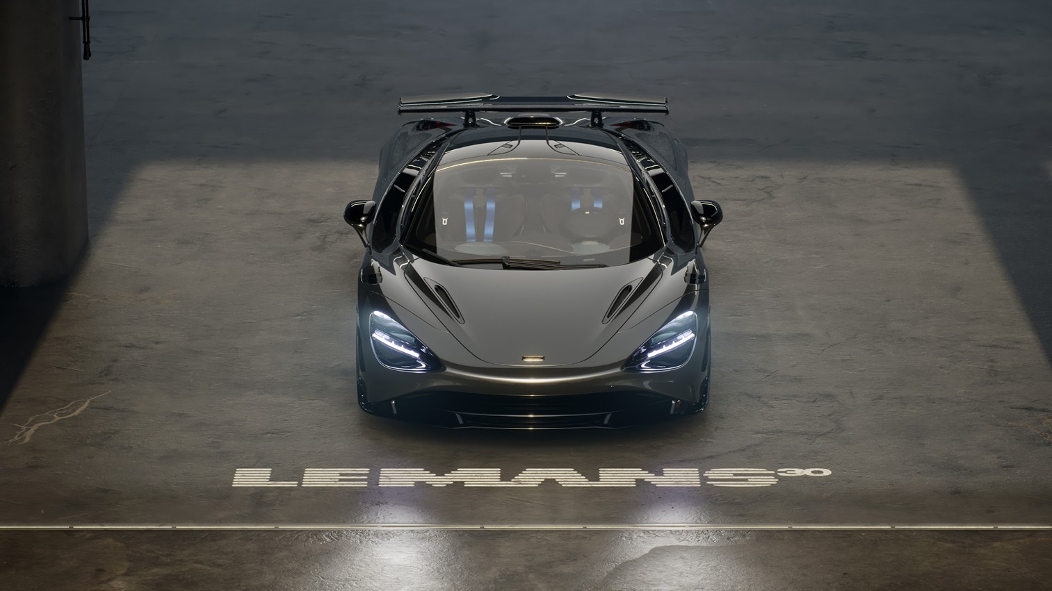 An aerial top-down view of a dark grey McLaren 750S Le Mans special edition, showcasing its sleek design, open-air cockpit, and the 'Le Mans 30' logo projected on the floor.