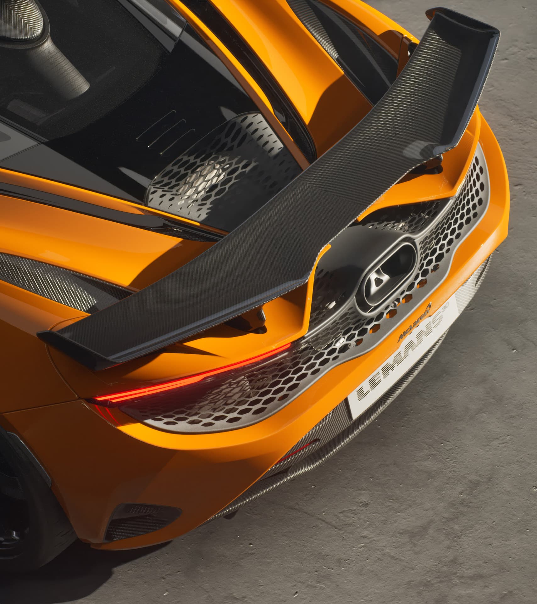 A close-up, top-down view of an orange McLaren 750S Le Mans special edition, highlighting its large carbon fiber rear spoiler, central exhaust, and honeycomb grille.