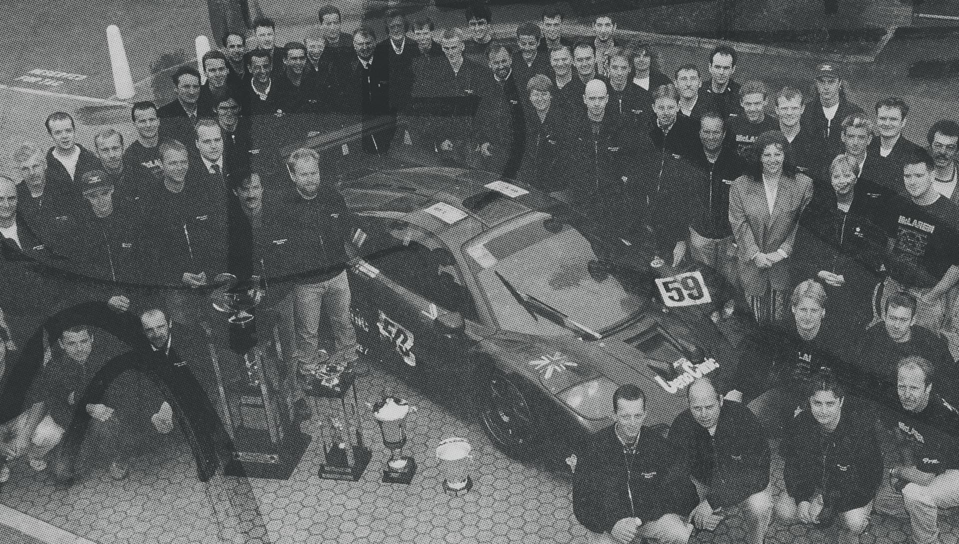 A vintage black and white photo of the McLaren F1 GTR racing team posing for a group photo with the number 59 race car and their trophy, celebrating their Le Mans victory.