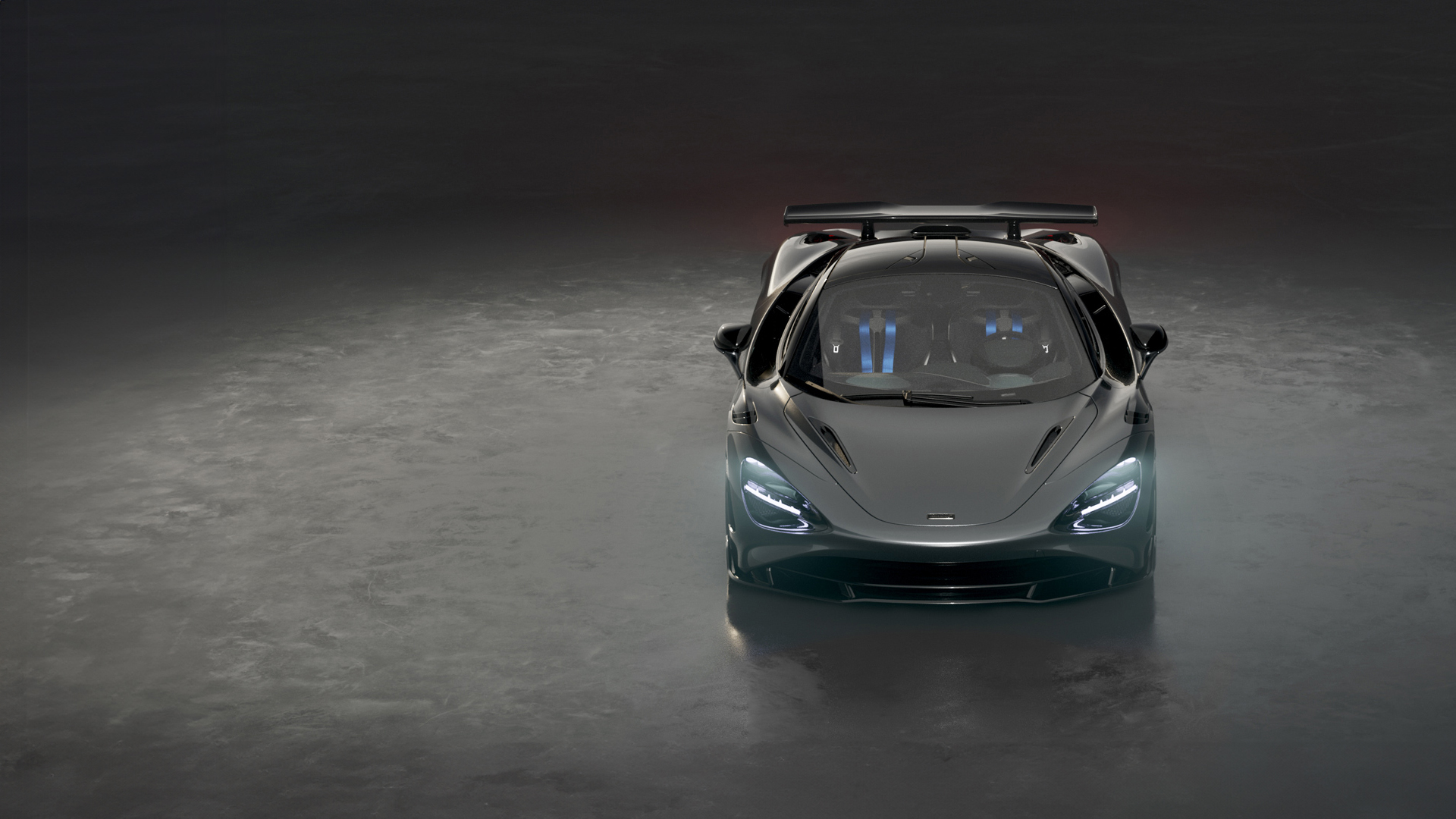 A dark grey McLaren 750S Le Mans special edition supercar is shown from a front-on view in a dark studio, with its headlights on and a prominent rear spoiler.
