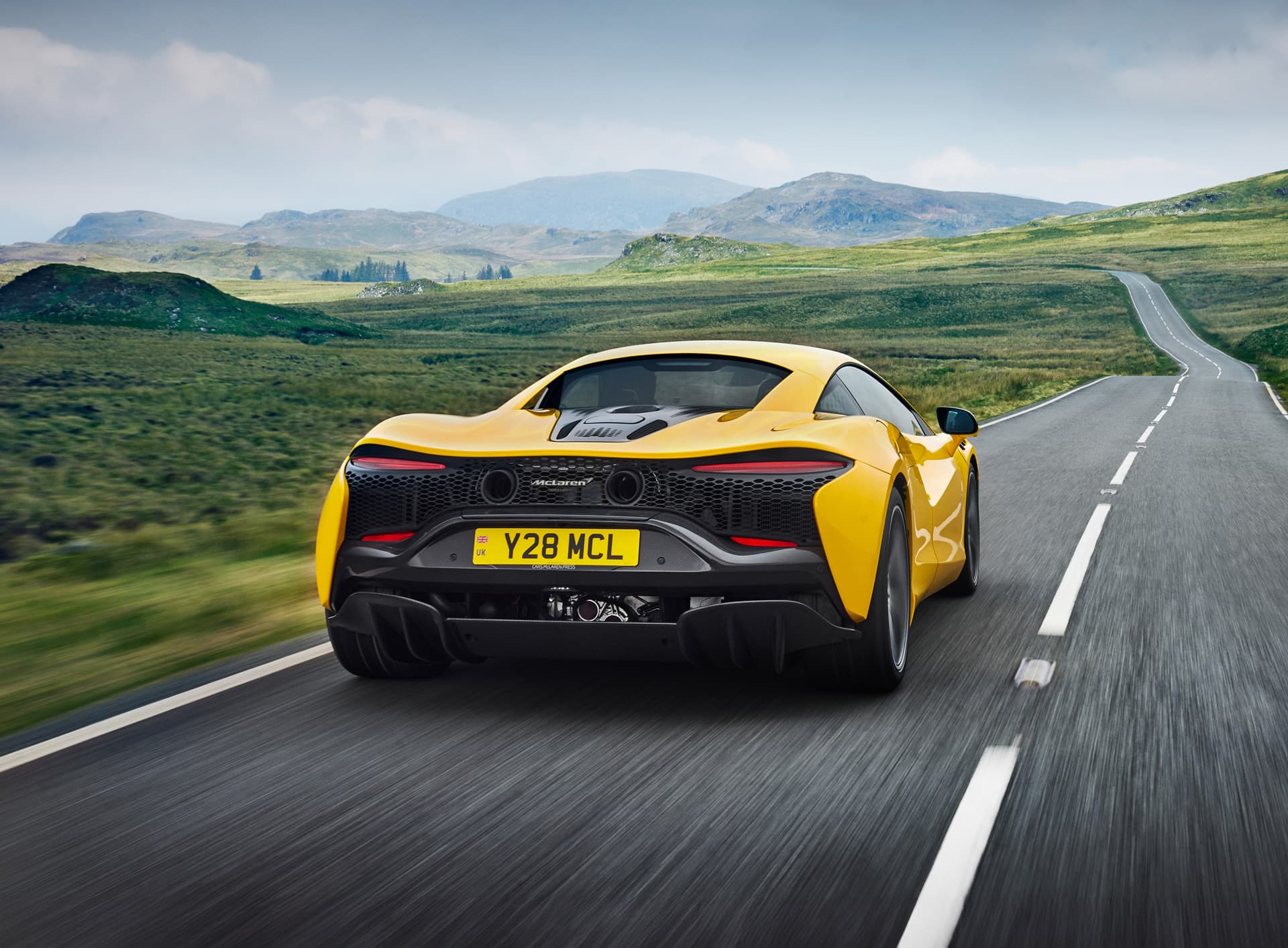  A rear-three-quarter view of a yellow McLaren Artura driving away on a winding road through green, rolling hills, showcasing its taillights and rear design.