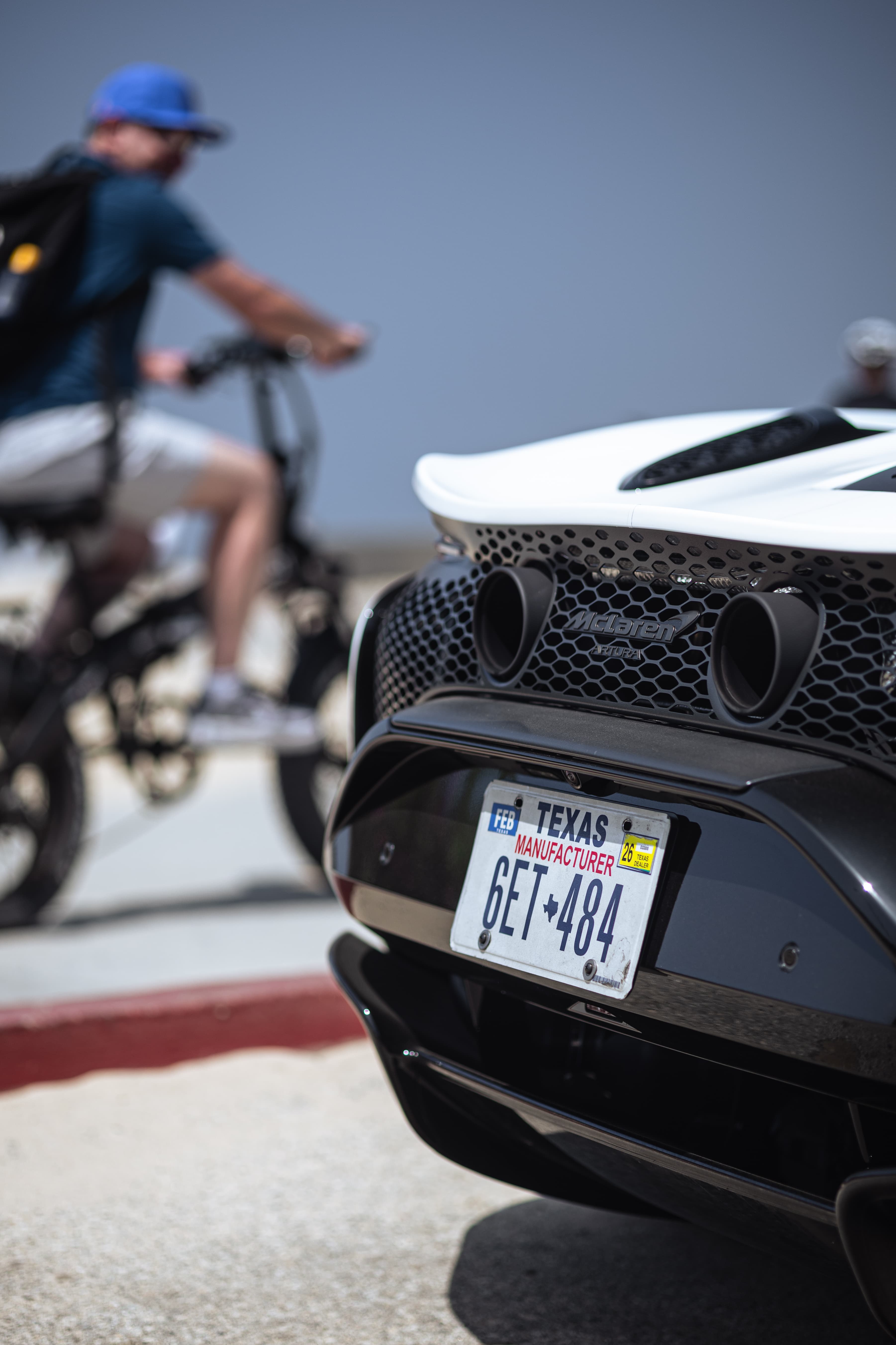 A close-up rear view of a white McLaren supercar with a Texas license plate and a dual exhaust, with a person on a bicycle blurred in the background.