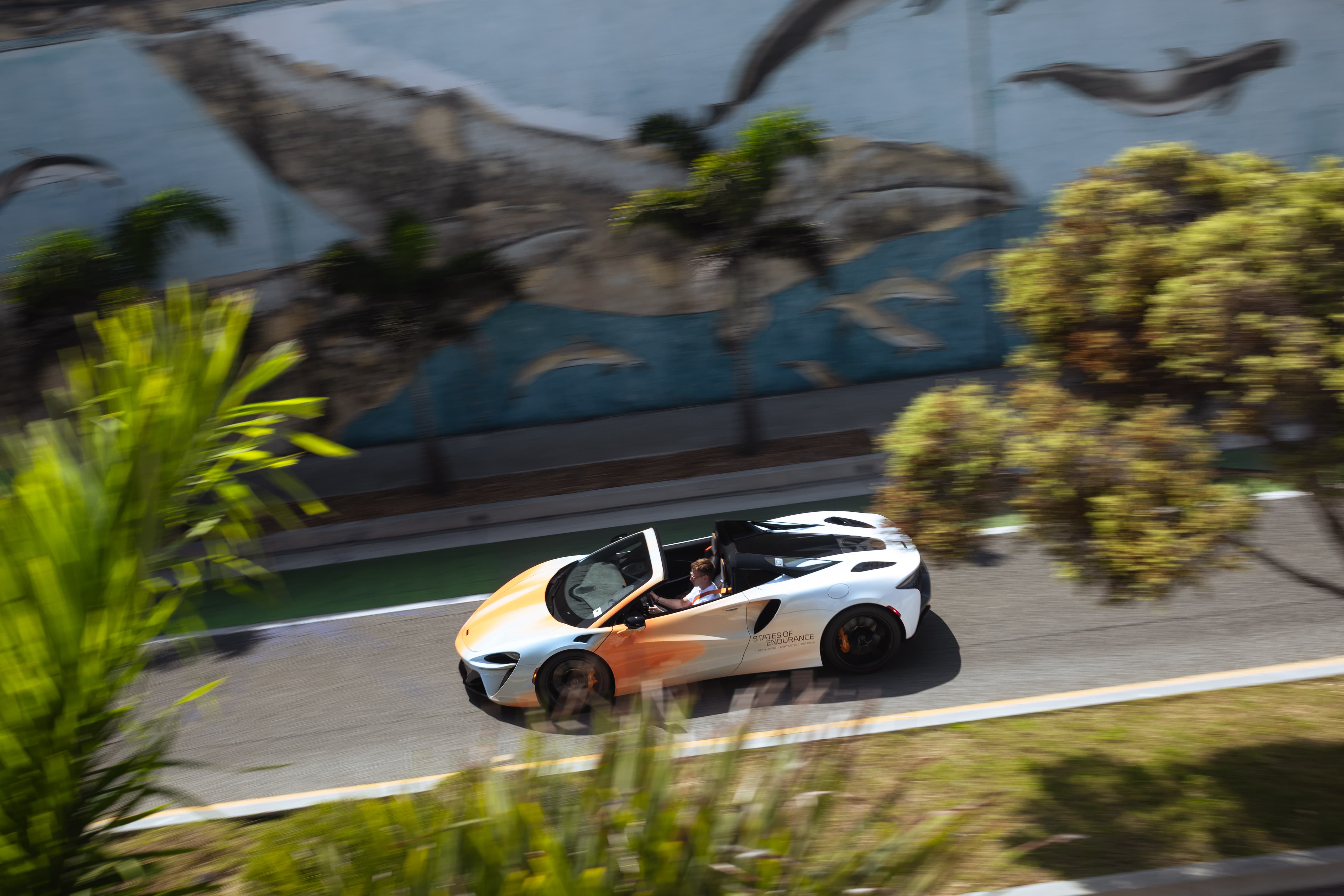 A dynamic action shot of a white and orange McLaren Artura Spider convertible driving at speed on a road, with a large marine-themed mural in the background.