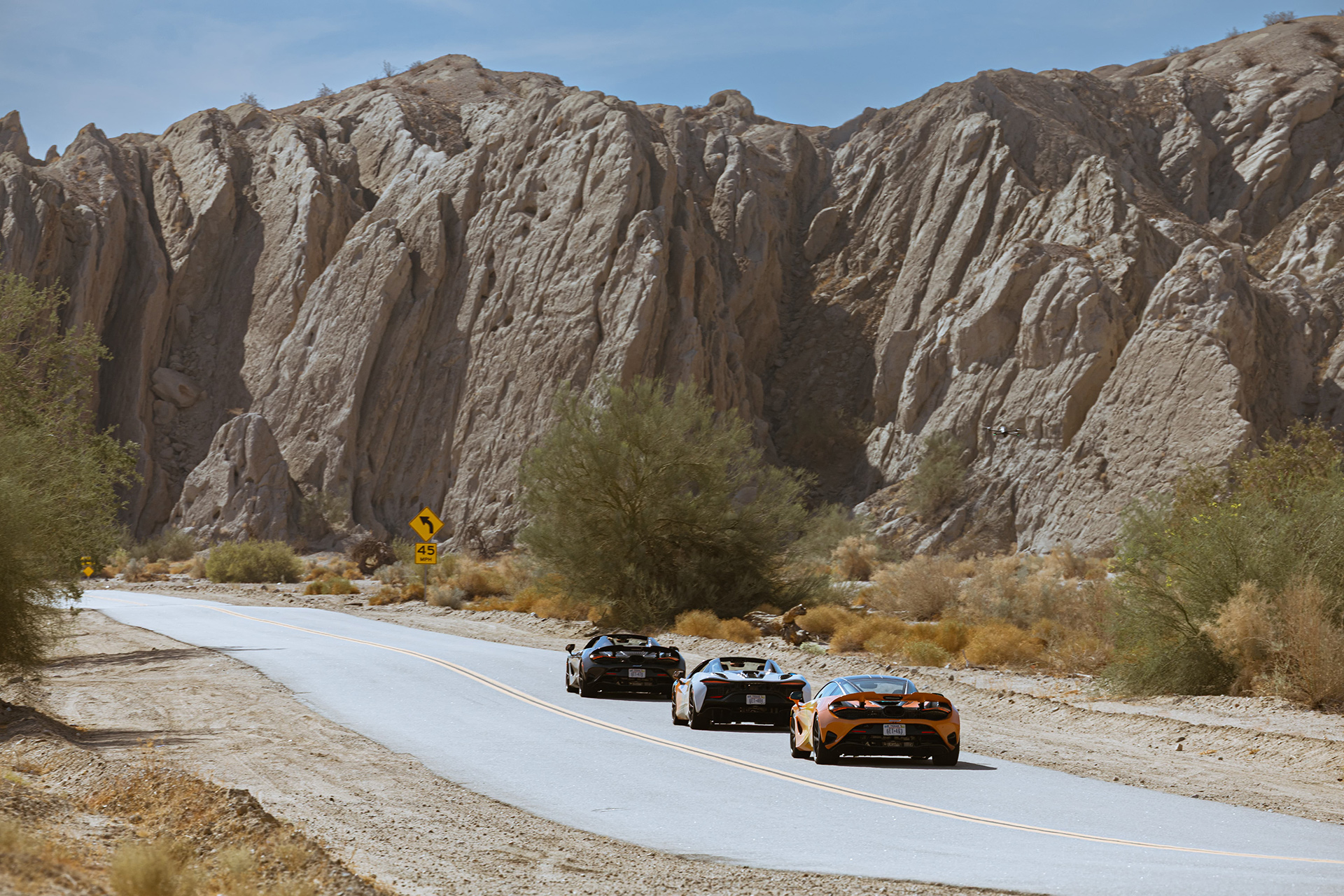 Three McLaren supercars with custom liveries are driving on a winding road through a desert landscape with rocky cliffs, part of the 'States of Endurance' campaign.
