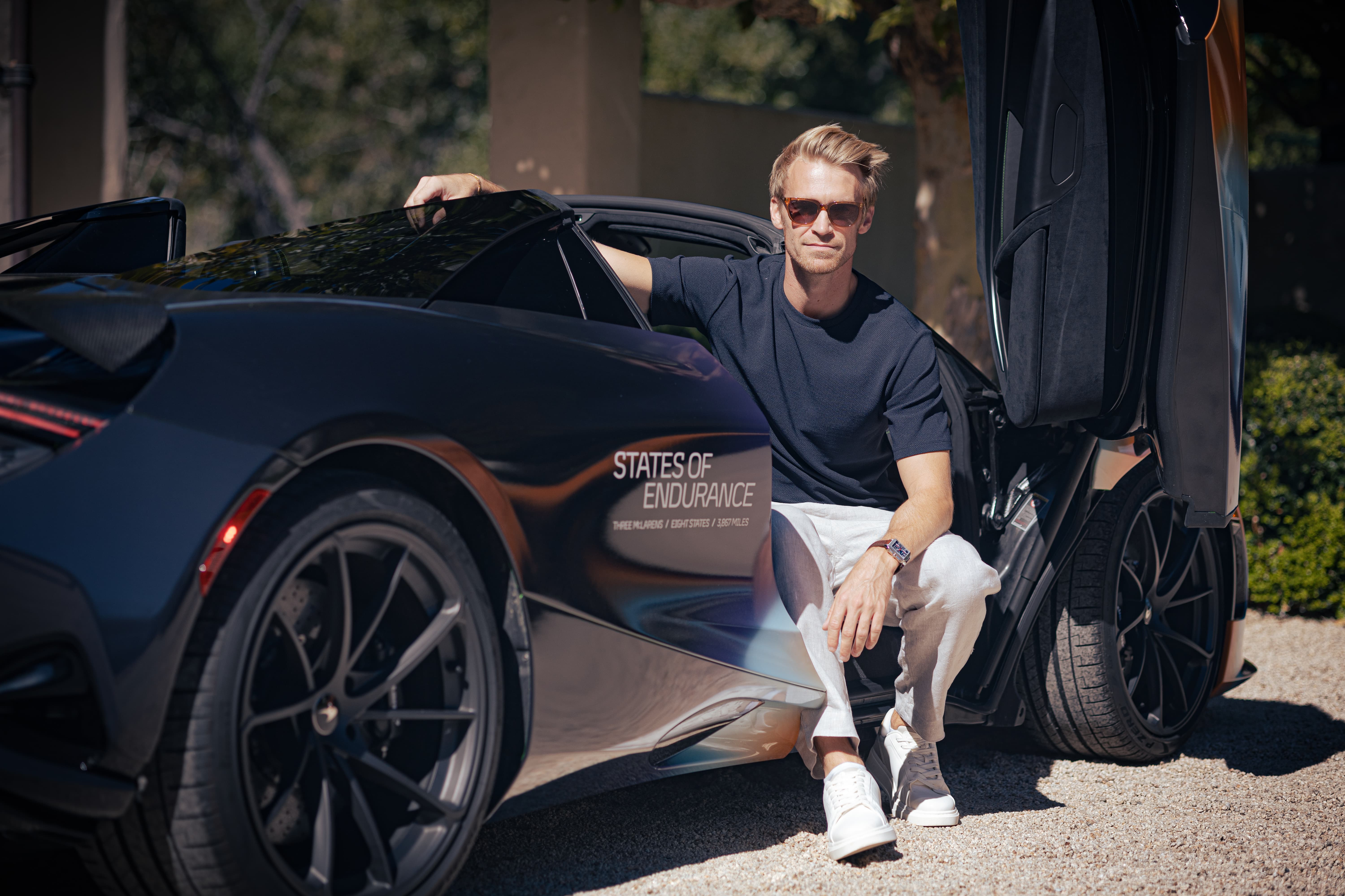 A man sits on the side of a custom-liveried black and purple McLaren supercar with its door open, highlighting the 'States of Endurance' logo on the body.