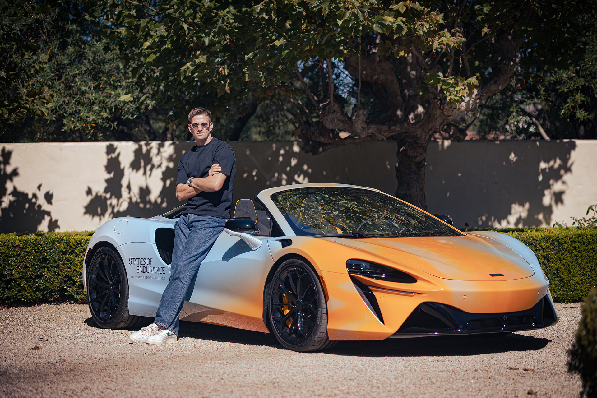 A man leans against a McLaren Artura Spider with a custom white and orange livery, in a group photo from the 'States of Endurance' road trip.