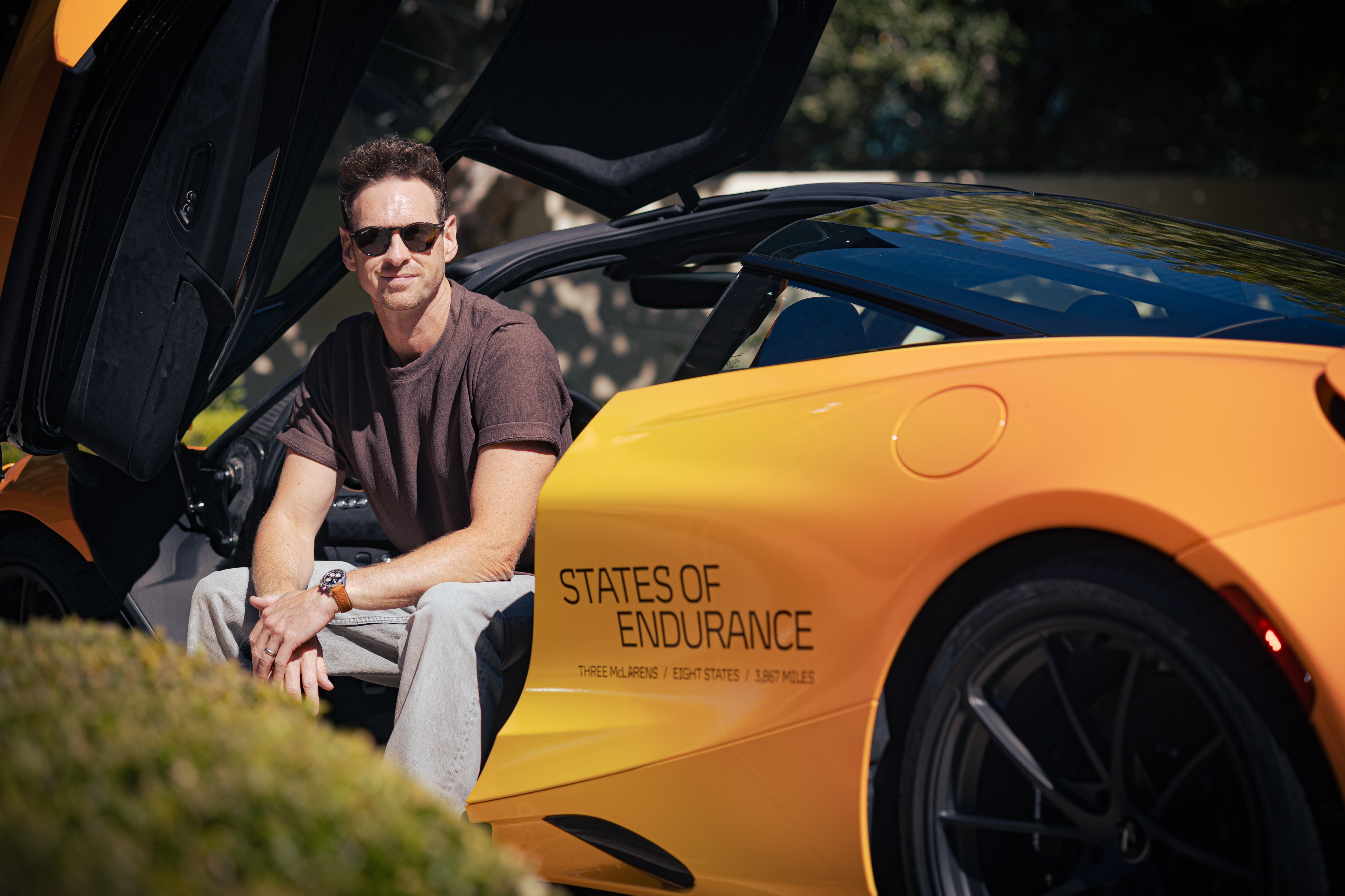 A man wearing sunglasses sits in the open driver's seat of a custom orange McLaren, showcasing the 'States of Endurance' logo and the journey's details on the side of the car.
