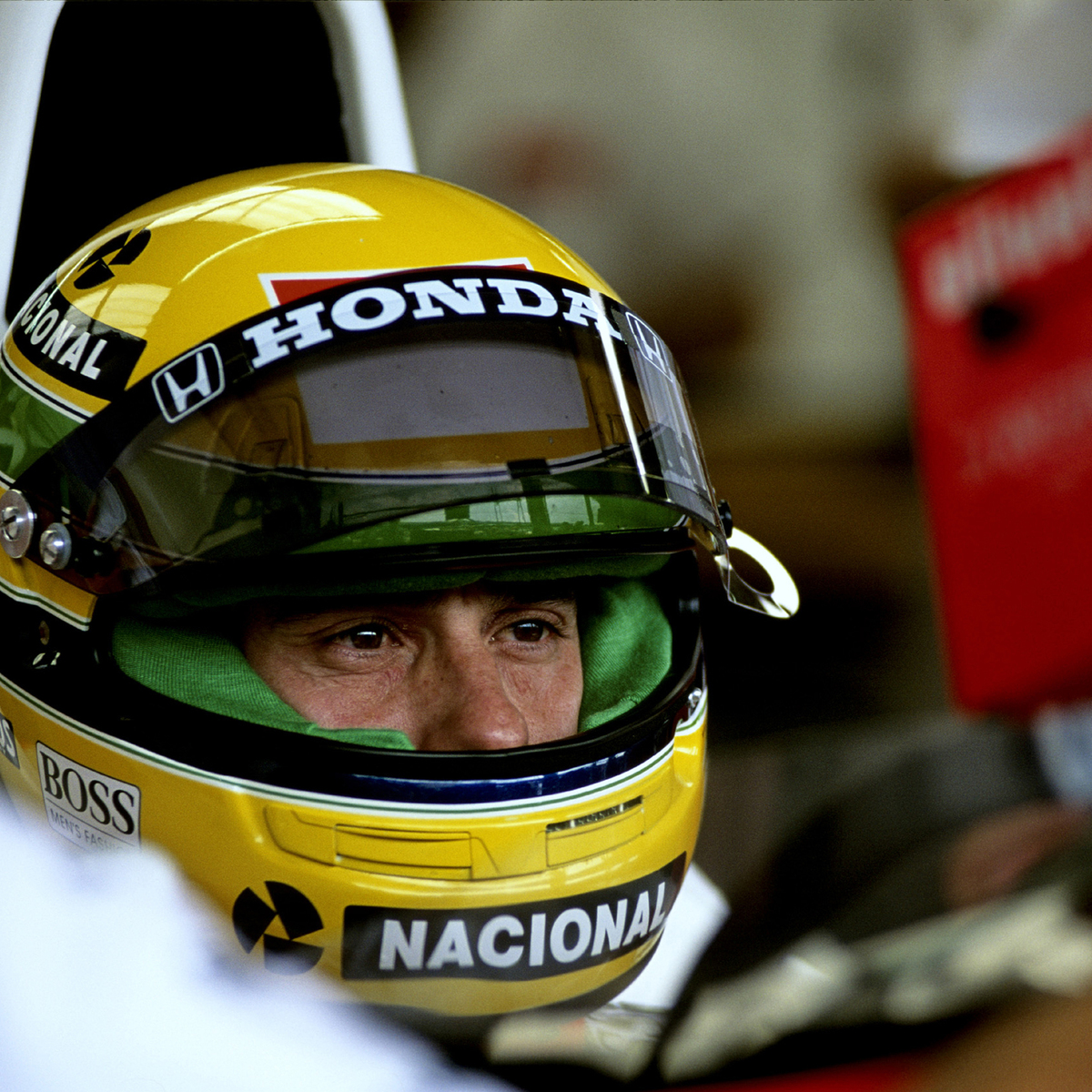 Ayrton Senna wearing his iconic yellow and green Formula 1 racing helmet with the visor up, so his eyes are visible.