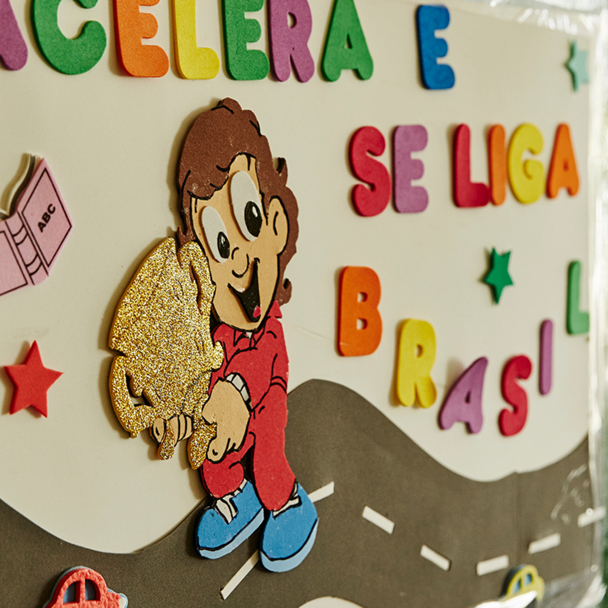 An 2D cartoon of Ayrton Senna holding a trophy made of foam, surrounded by Spanish text and stars made of felt, on a paper display board in a school. 