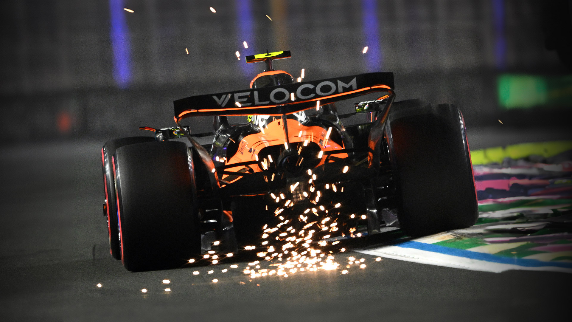 The rear of the McLaren MCL39 F1 car kicking up a shower of sparks at high speed on the track at night.