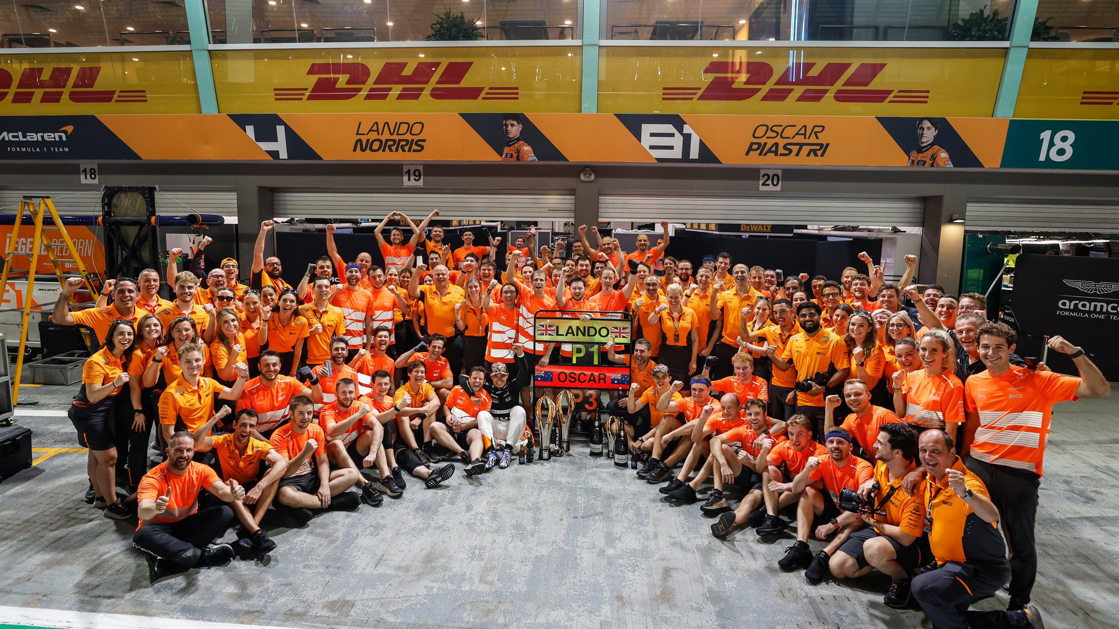 A celebratory group photo of the McLaren Formula 1 team and crew in their orange uniforms, standing in a pit lane with their drivers' names in the background.