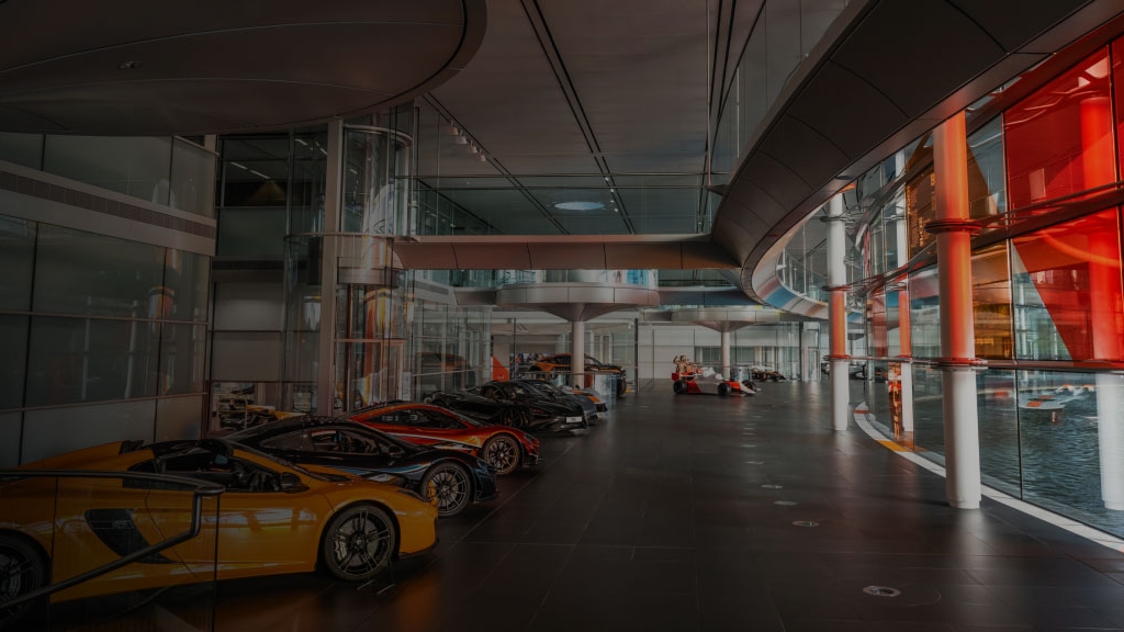 A spacious, modern hall featuring a row of parked McLaren supercars in various colors (yellow, black, red, blue) on the left, and a large glass wall on the right reflecting a red structure and water.