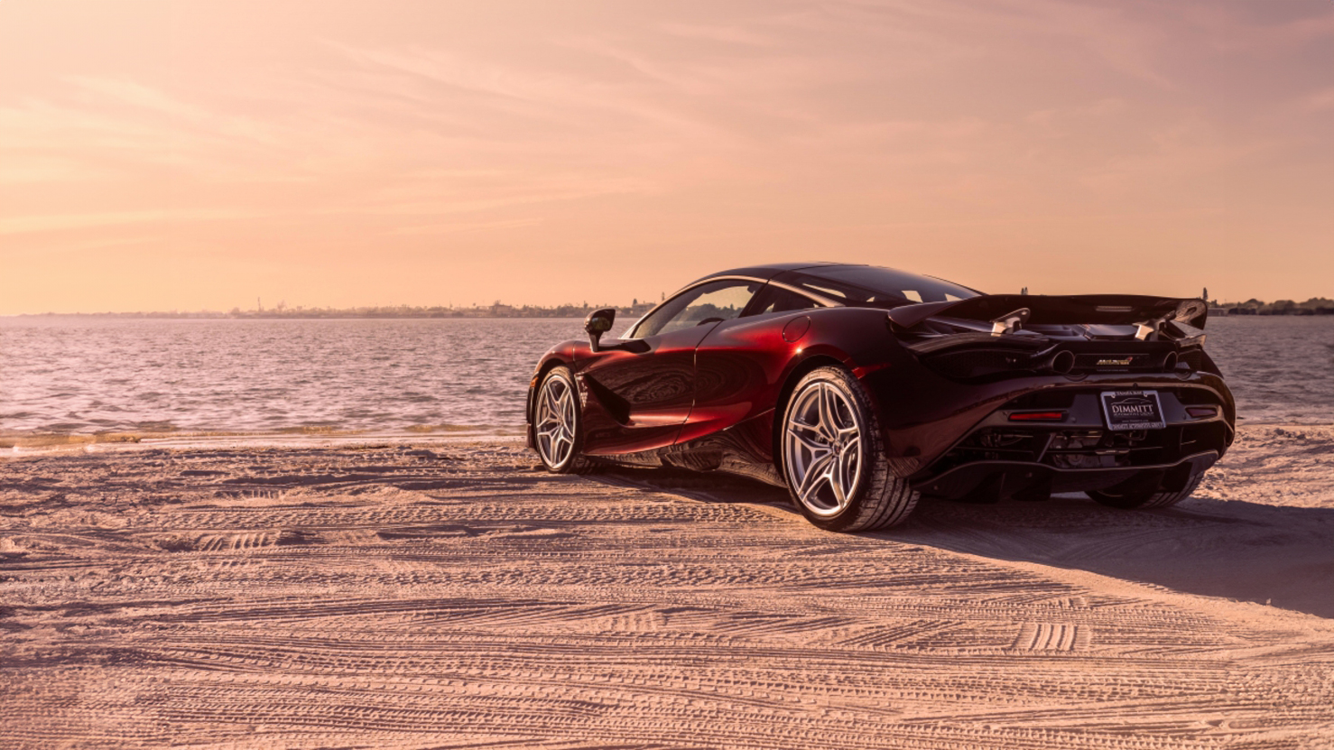 A stunning red McLaren 720S sports car is parked on a sandy beach next to the ocean at sunset, with a vibrant orange and pink sky.