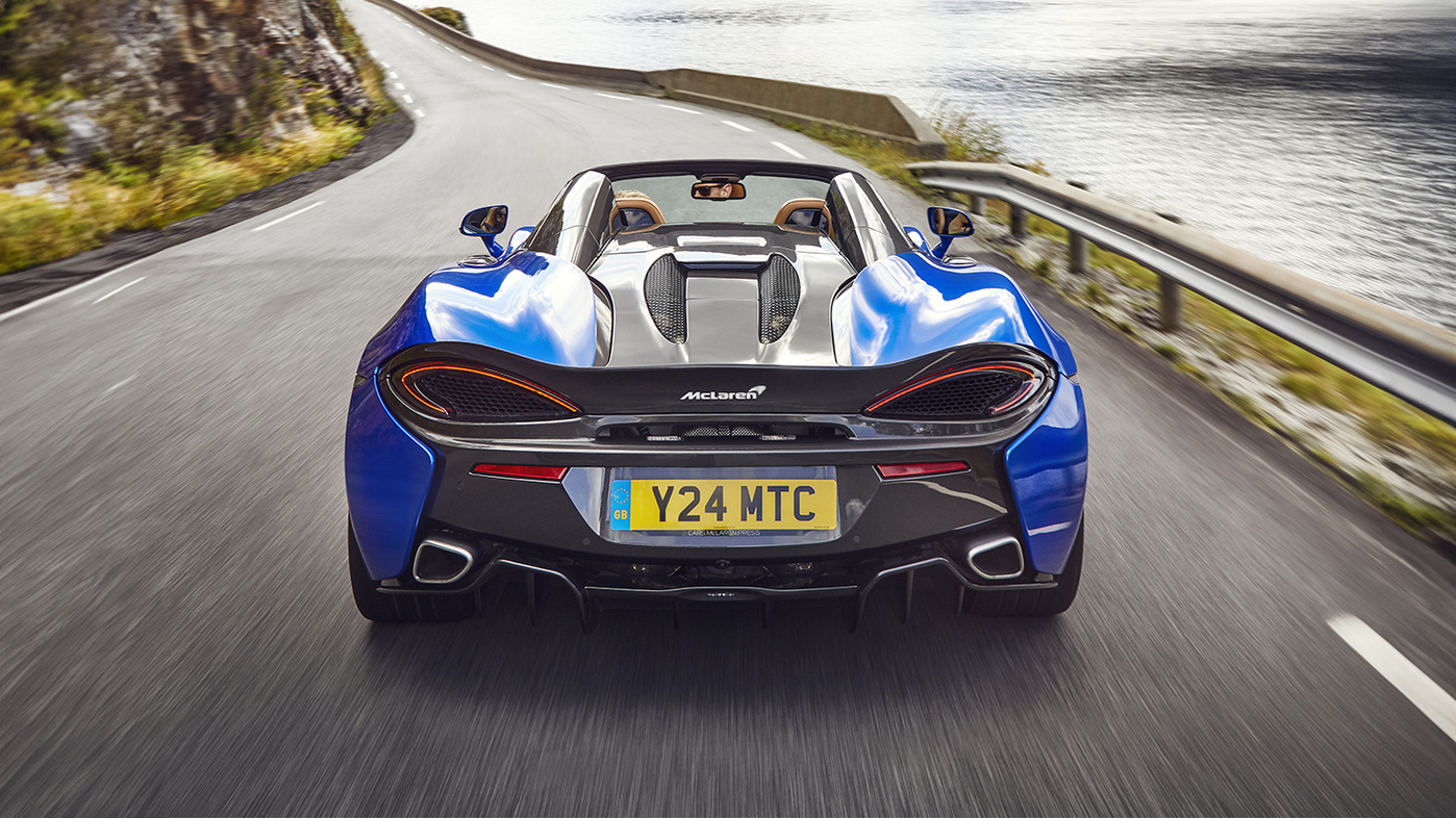 A rear-view action shot of a blue McLaren 570S Spider convertible driving on a road next to a large lake with mountains in the background.