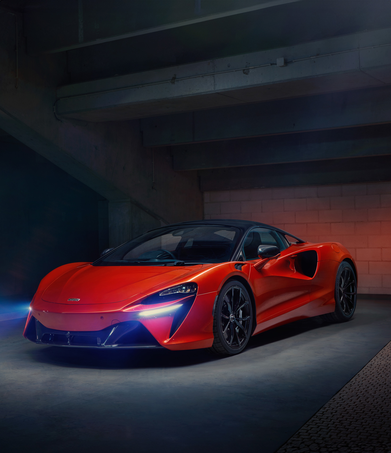 The McLaren Artura hybrid supercar in a vibrant red finish, parked in a dimly lit garage with its headlights glowing.