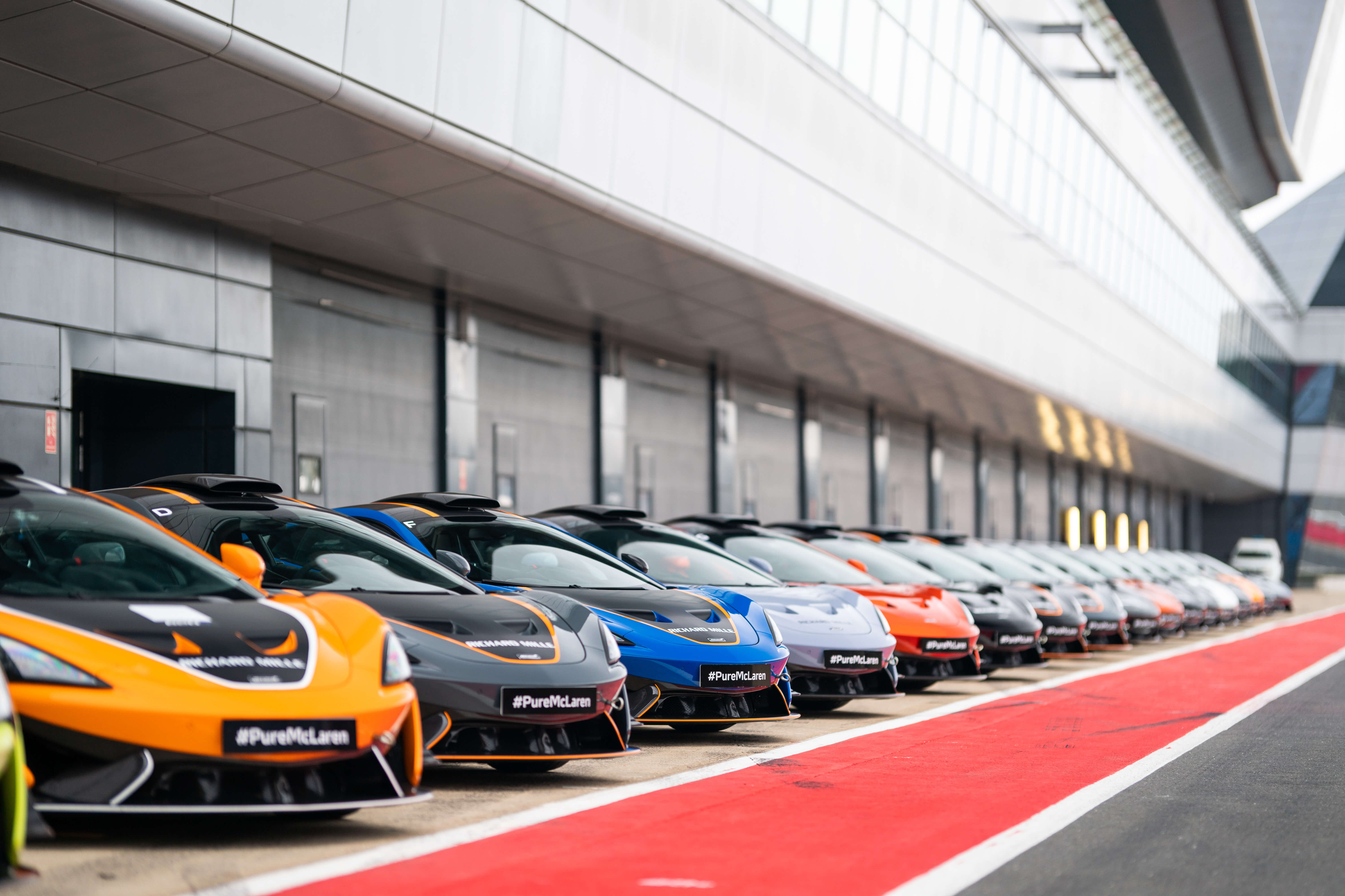 A long line of McLaren supercars in various colors, parked in a pit lane with "PureMcLaren" branding on the registration plate.