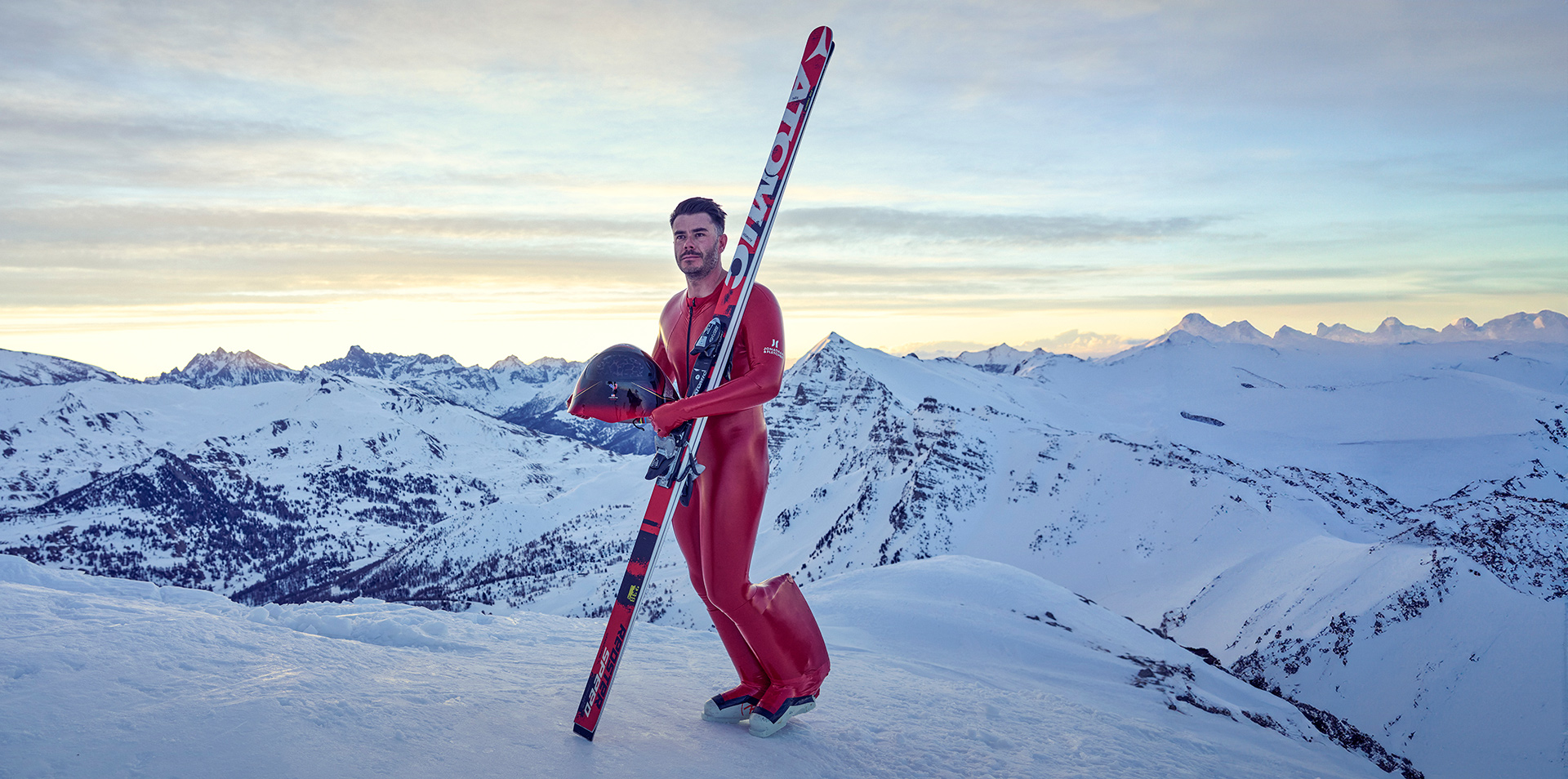 Simon Billy stood on a snowy mountain side, wearing full red ski gear, holding his skis and helmet. 