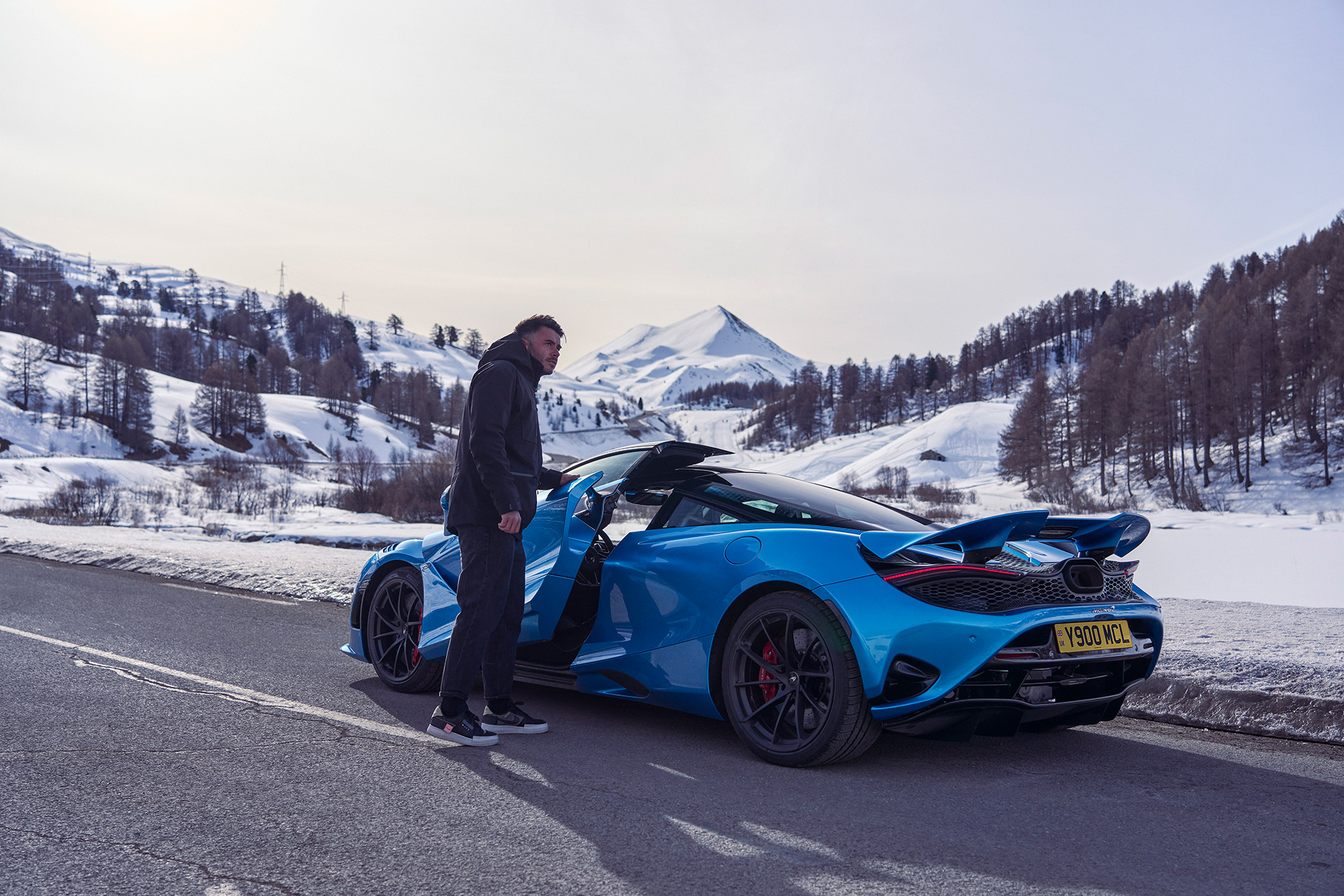 Simon Billy opening the door of a bright blue McLaren 750S, parked on a snowy mountain road.