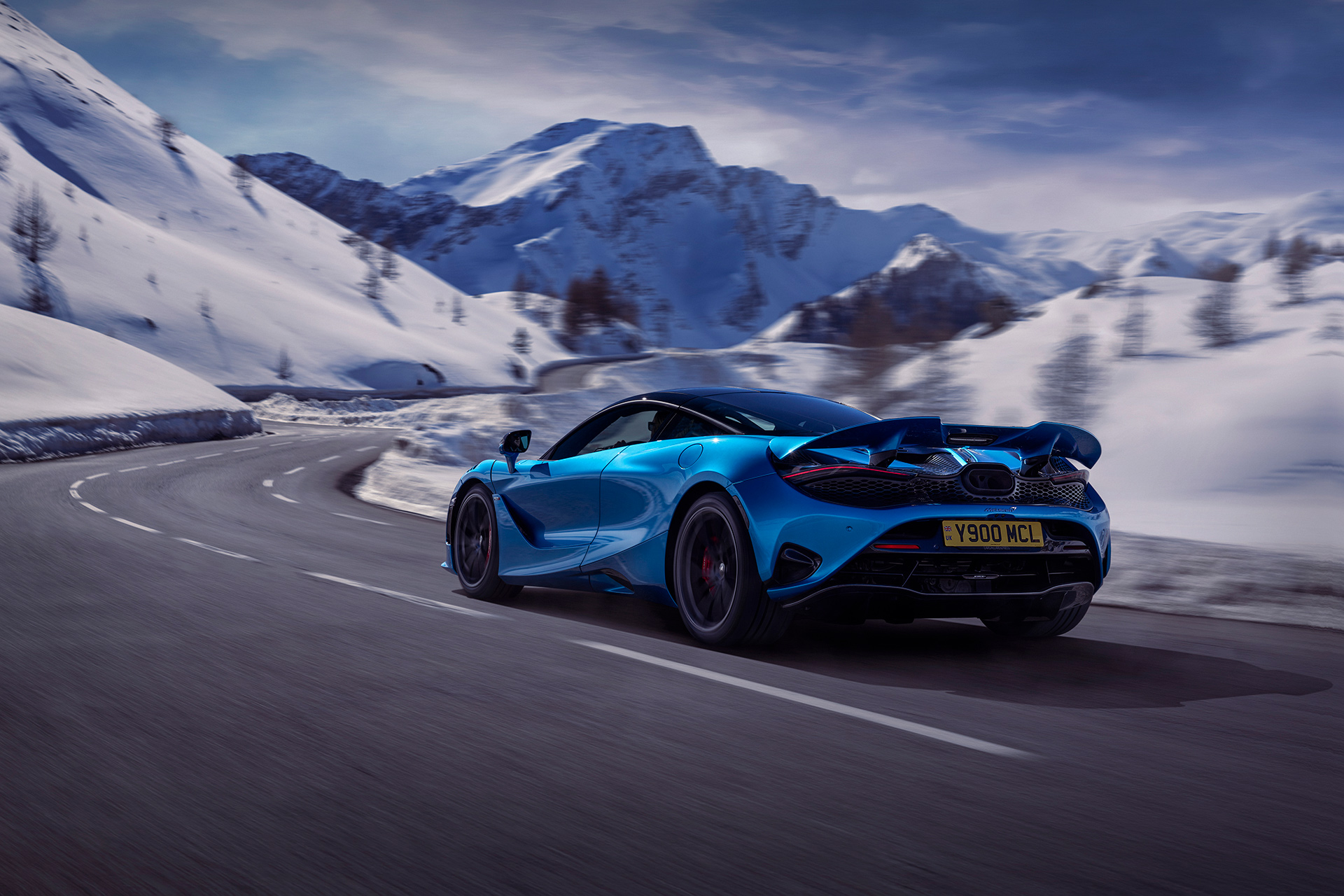Rear side view of a Simon Billy driving a blue McLaren 750S down a snowy mountain road at speed.