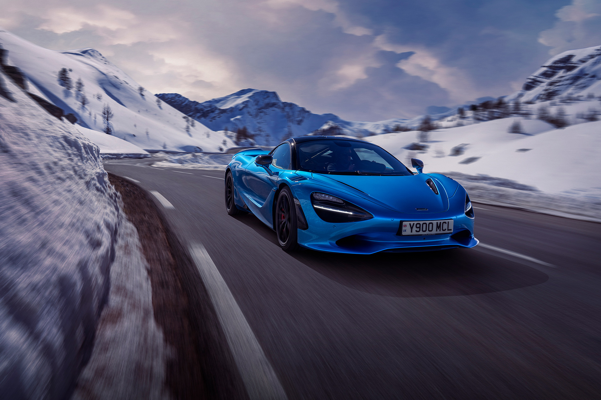 Front side view of a Simon Billy driving a blue McLaren 750S down a snowy mountain road at speed.