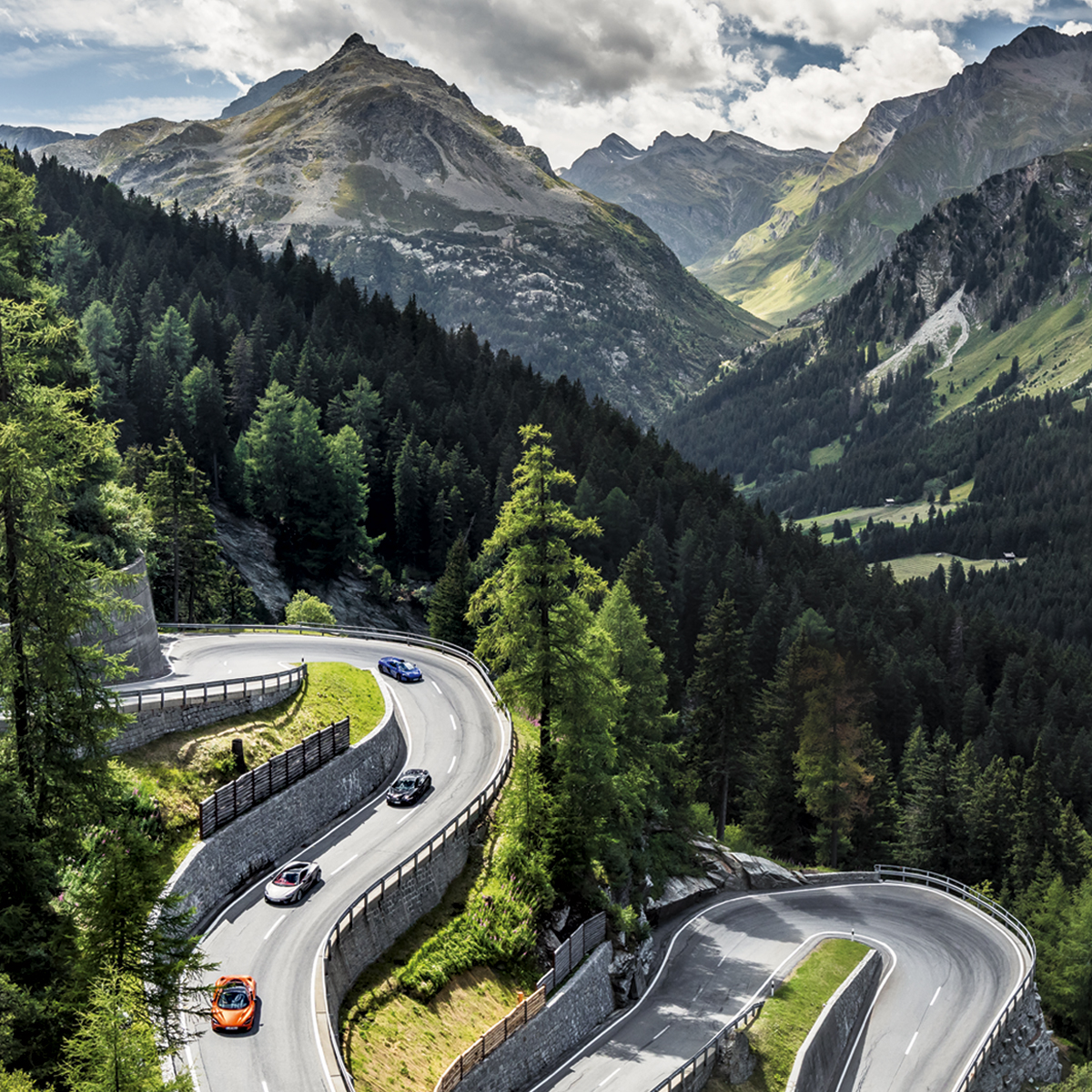 Series of McLaren cars driving down a winding mountain road, surrounded by trees.