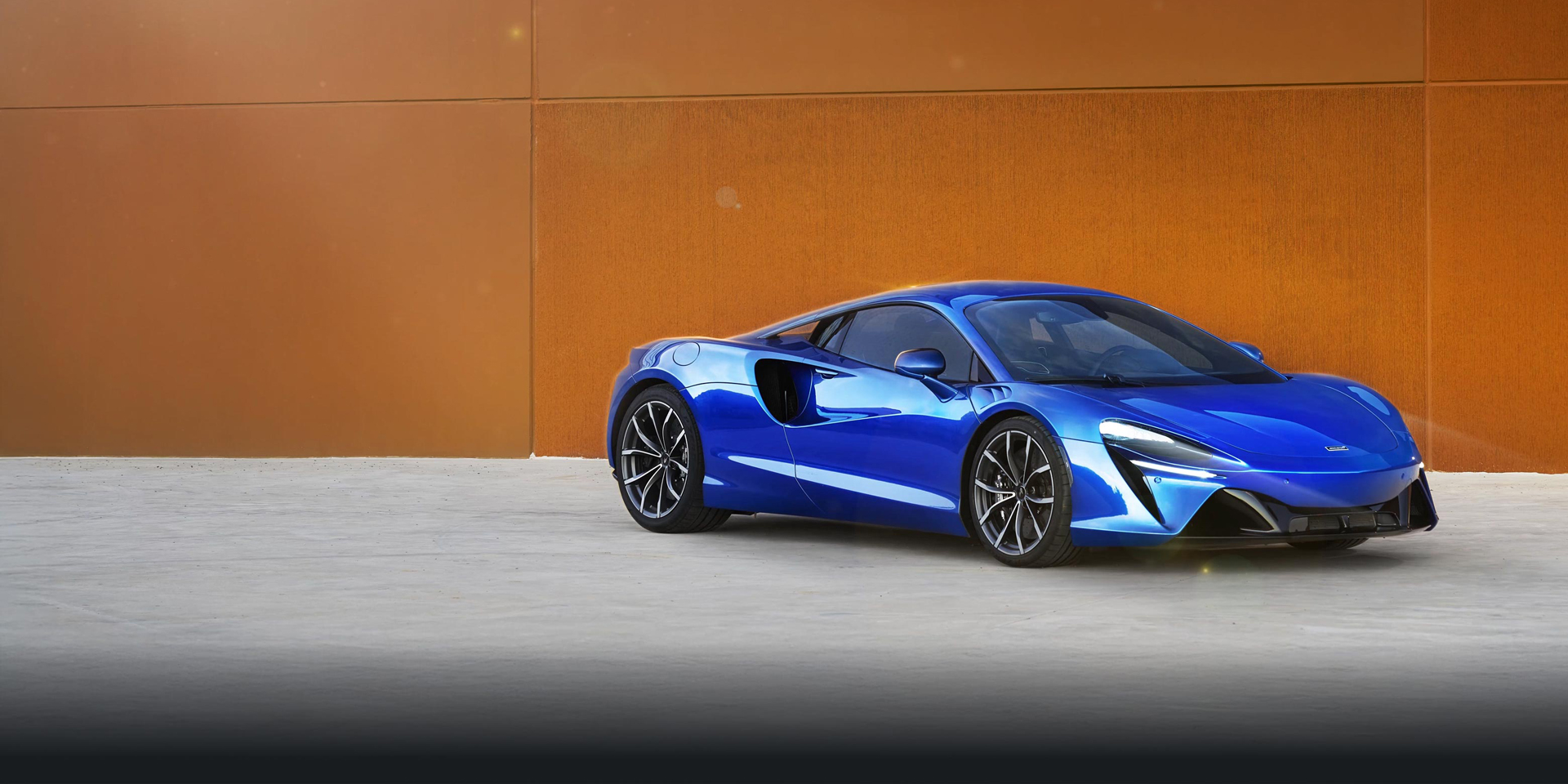 Blue McLaren Artura parked in front of an orange building in the sun.