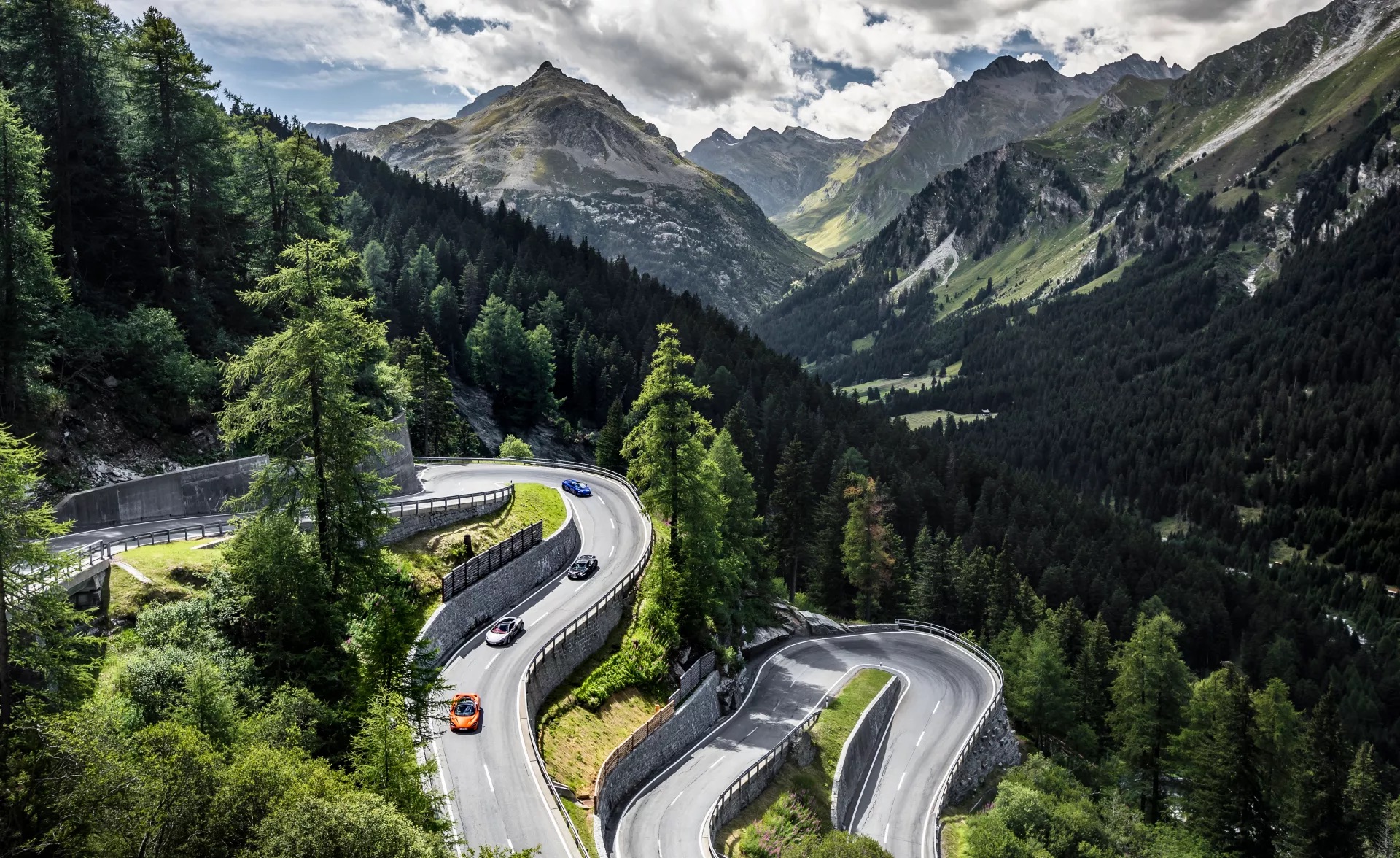 A convoy of McLaren sports cars in various colors navigating a dramatic, winding mountain pass, showcasing the ultimate grand touring experience.
