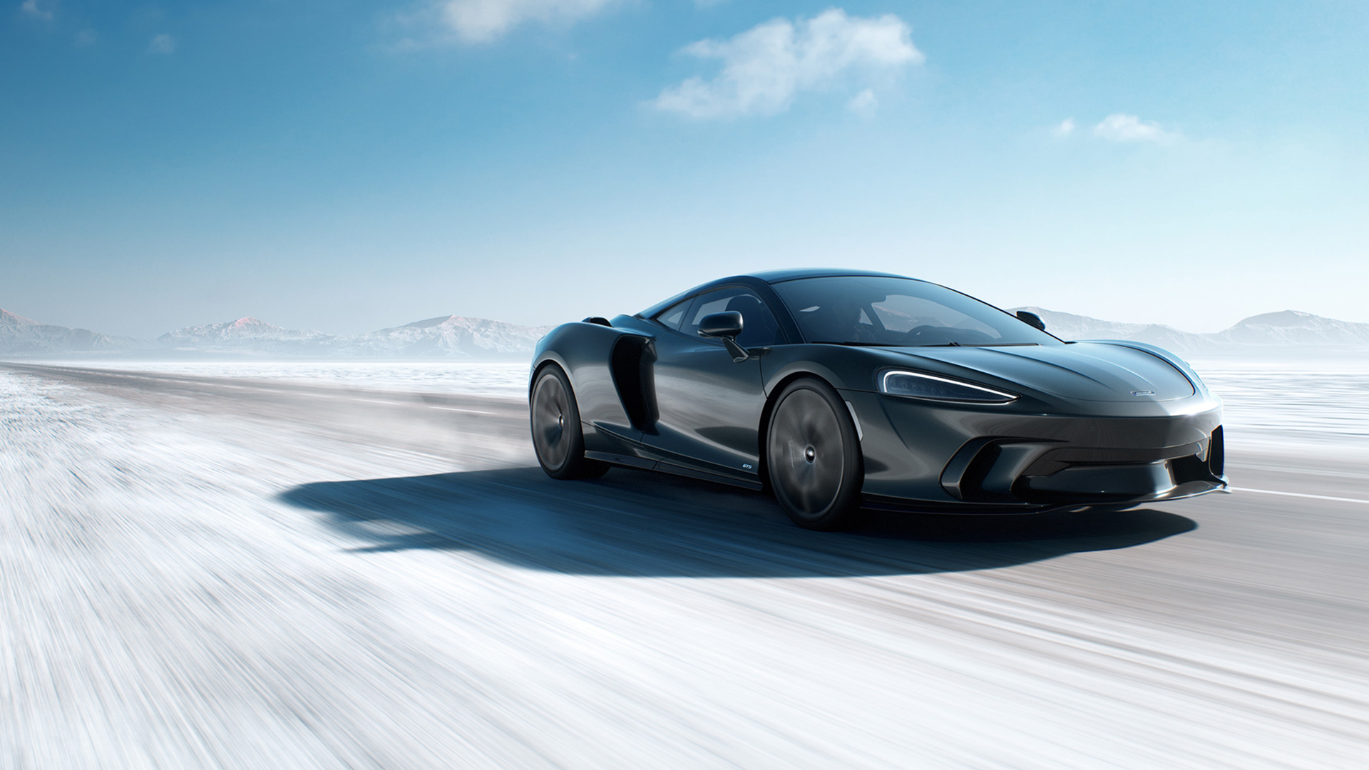 McLaren GTS high-performance sports car driving fast on a frozen, snow-dusted road. The sleek, dark grey GTS demonstrates its all-weather capability and precision-engineered power in the extreme winter landscape.