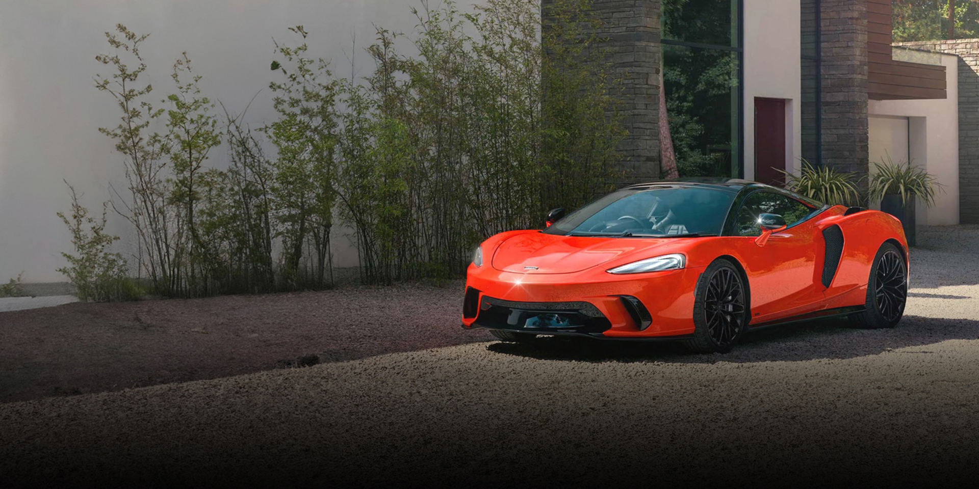 A bright red McLaren GTS is parked on a gravel driveway in front of a modern house with stone and white walls and large windows, surrounded by trees.