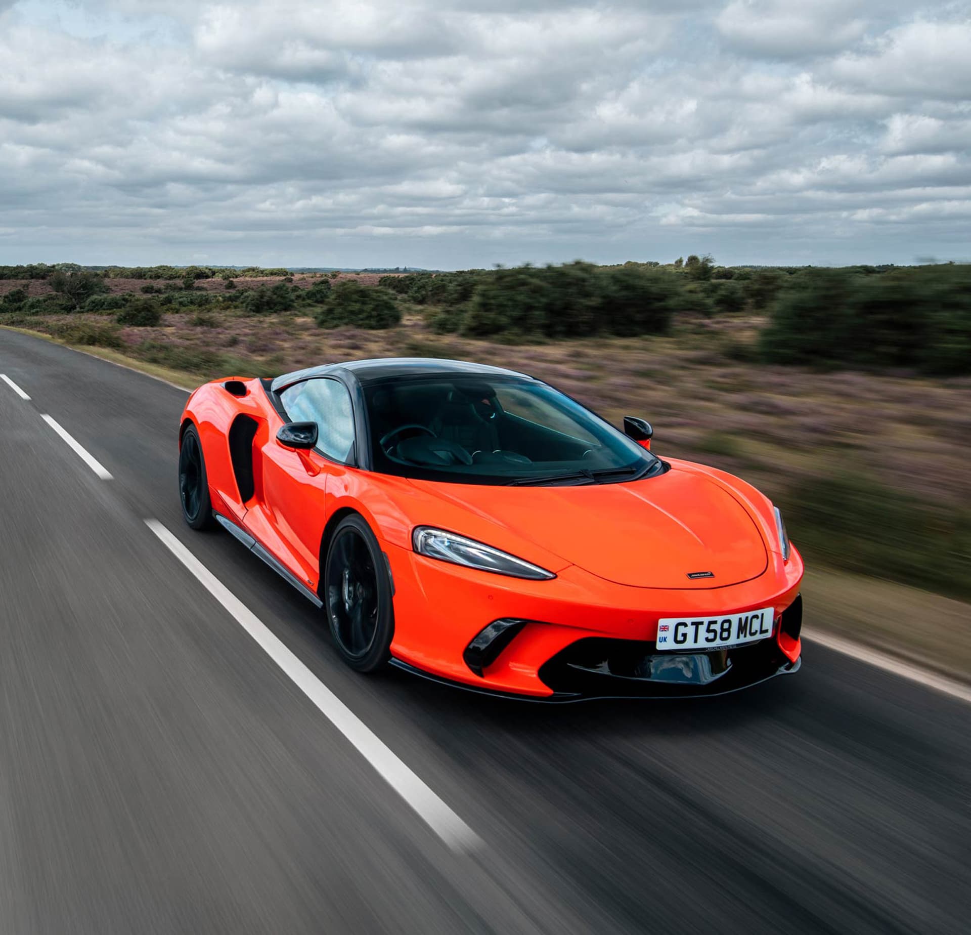 A bright red McLaren GTS speeds along a winding two-lane road in a vast, open landscape under a dramatic, cloudy sky, with blurred surroundings conveying motion.